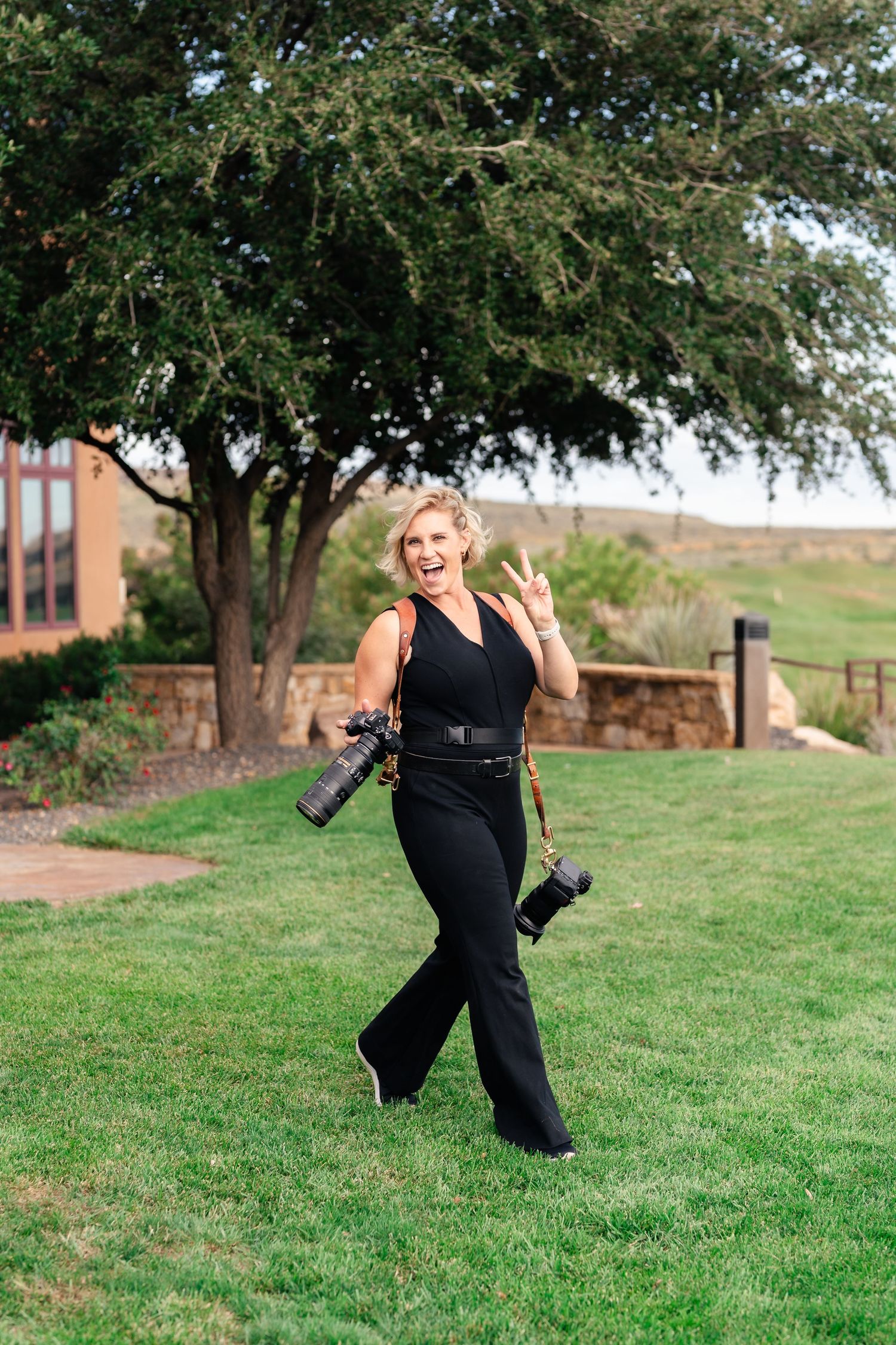 Professional photographer walking on grass holding two DSLR cameras while wearing a black jumpsuit outdoors.