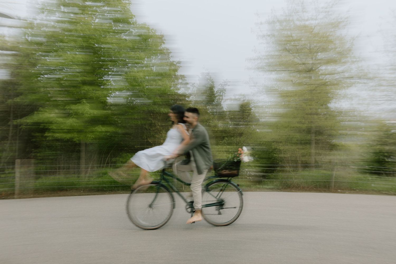 Cyclist rides by at high speed with camera capturing motion blur effect on a paved path surrounded by green trees.