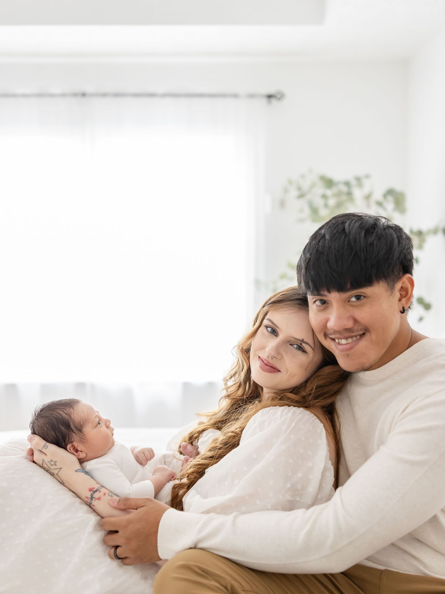 A couple in white sweaters cuddles on a bed with their newborn baby in a bright, airy bedroom.