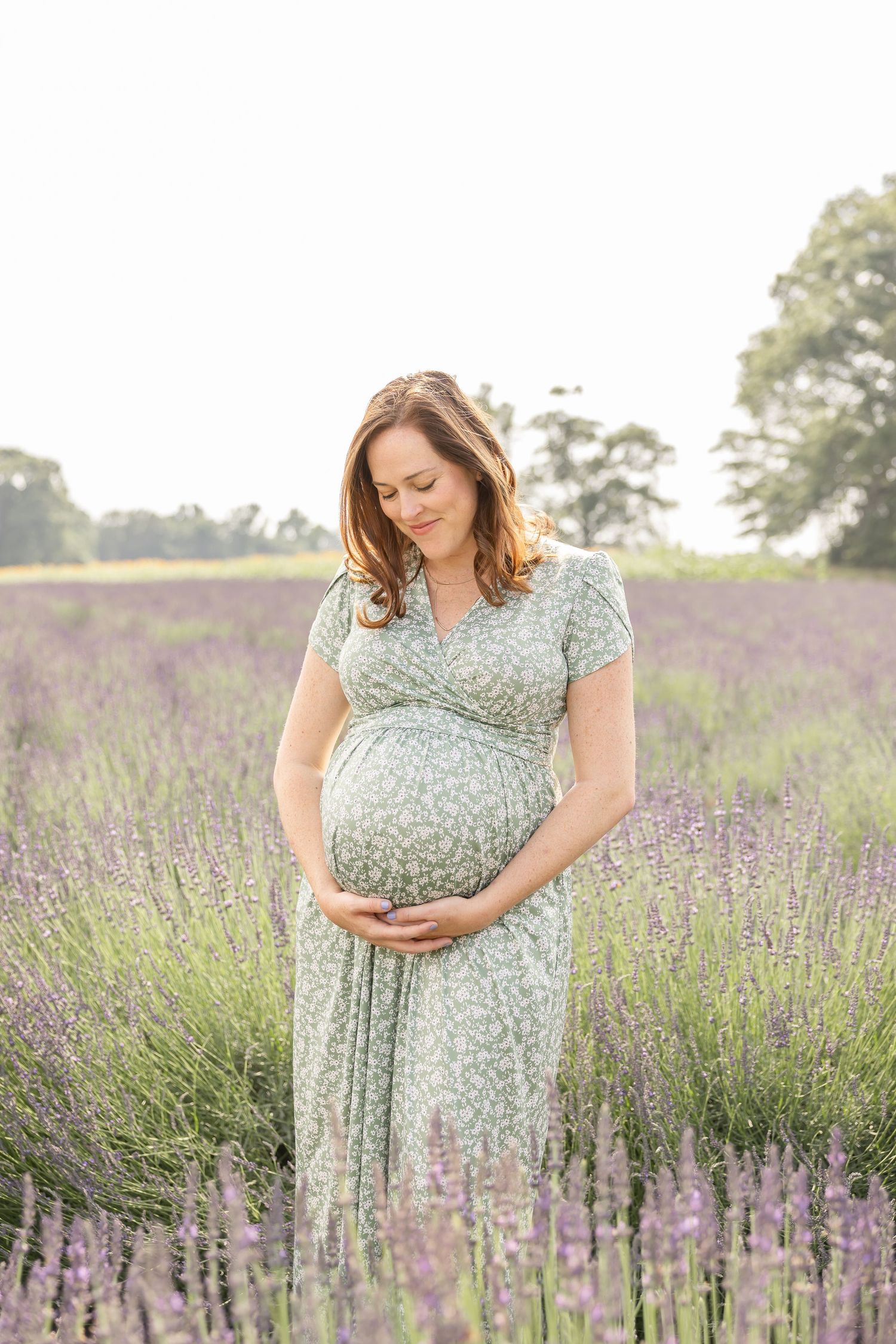 Expectant mother in a floral dress stands serenely in a blooming purple lavender field on a sunny summer day.