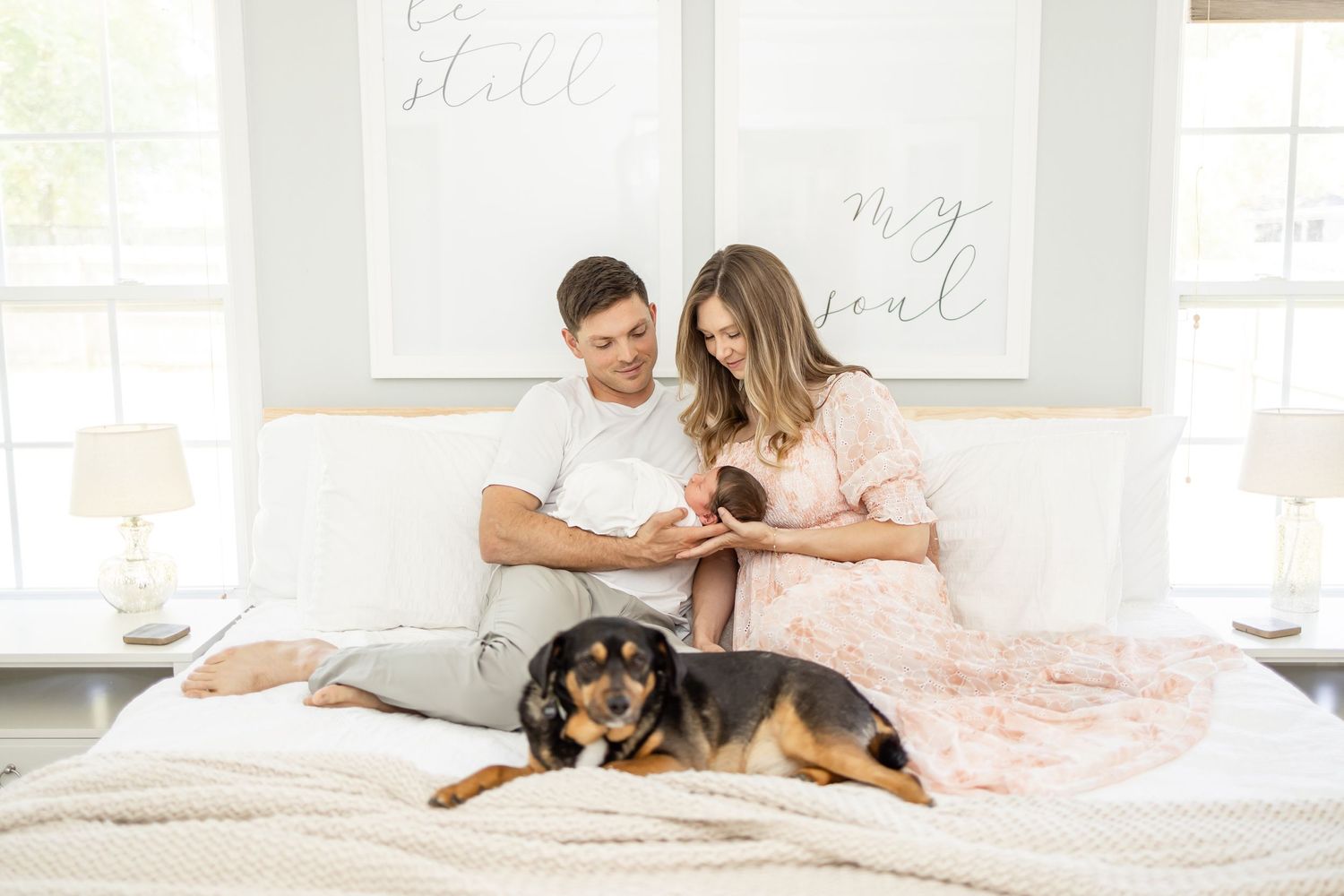 A couple relaxes in bed with their black and tan rescue dog laying at their feet on white bedding.