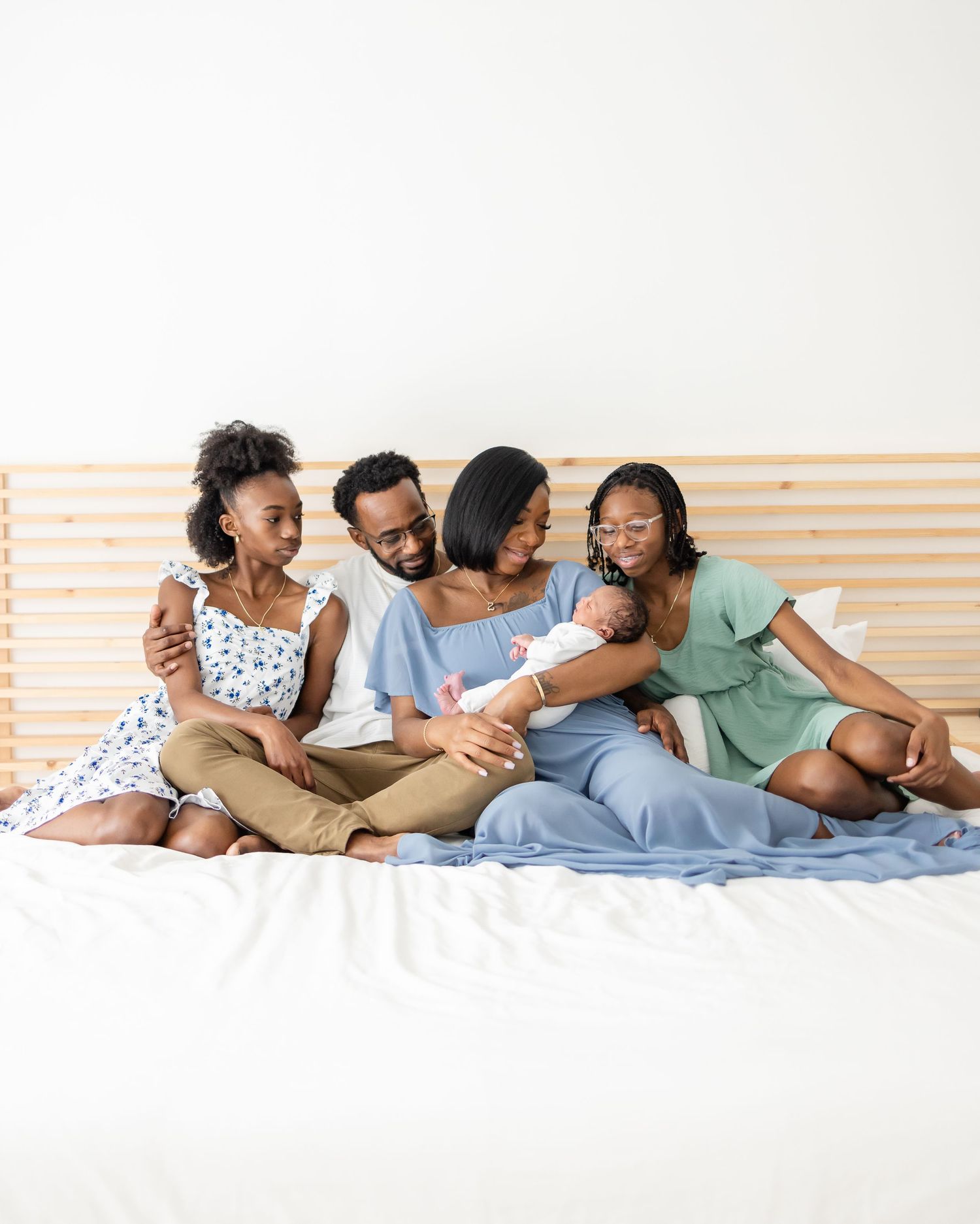 A family sits together joyfully on a bed with white bedding against a wall with light wooden slats.