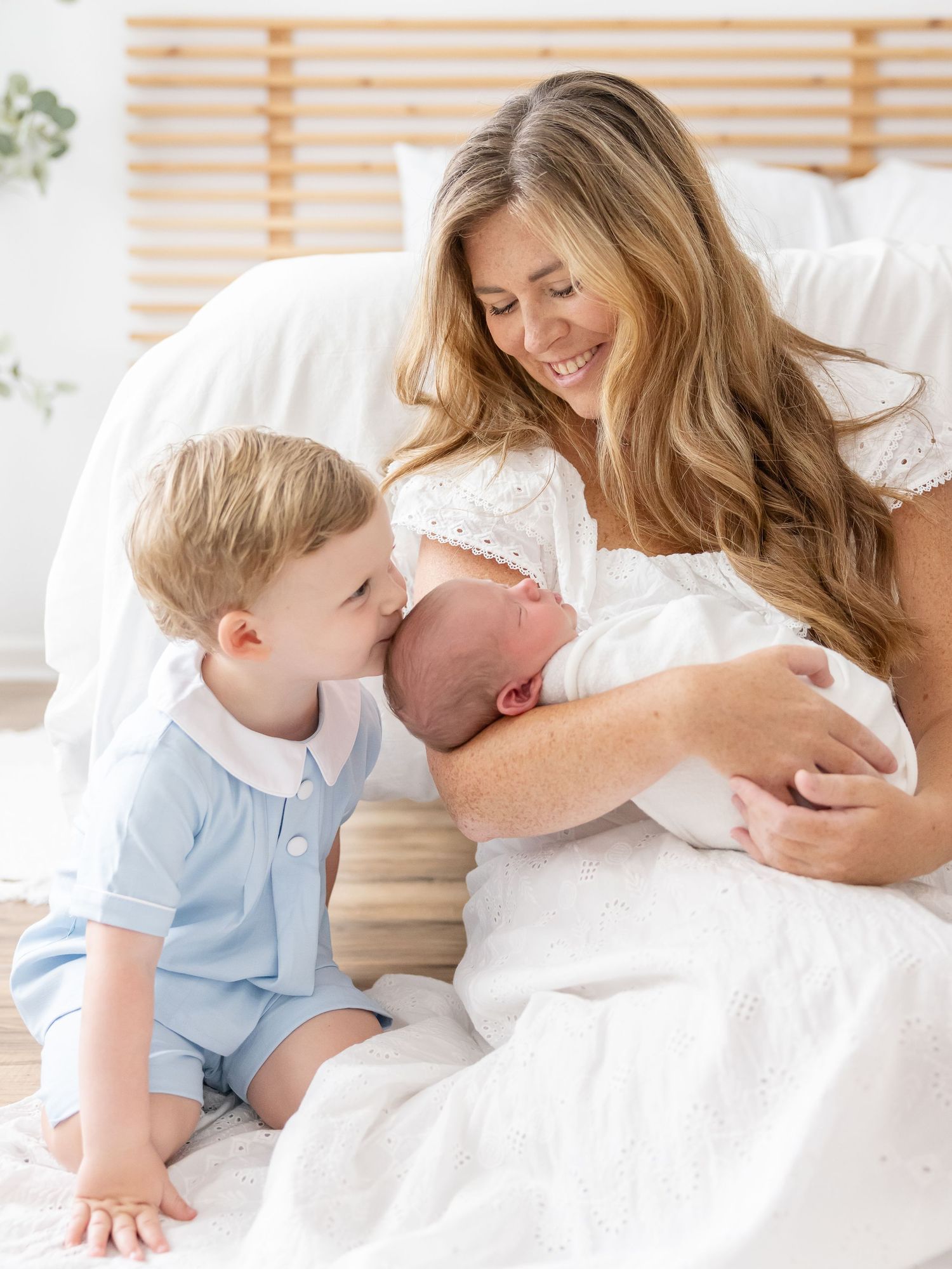 A tender moment as a mother in white holds her newborn baby while her young son in blue gently places a kiss on top of baby's head.