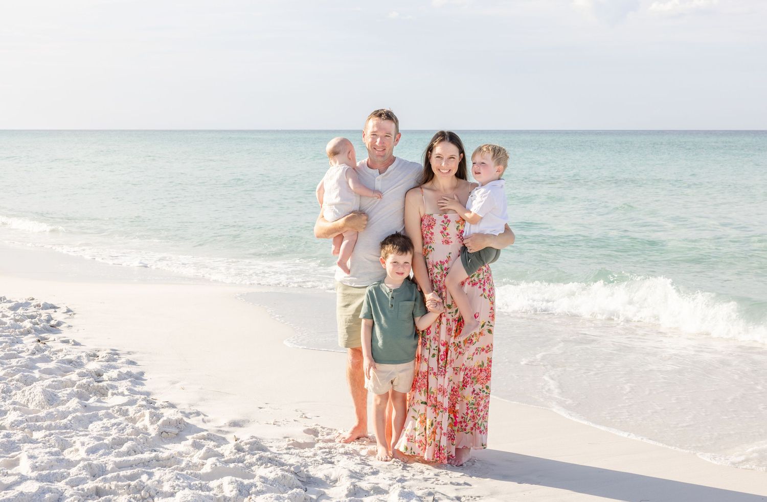 A family in summer attire poses together on a pristine white sand beach with turquoise ocean waves behind them.
