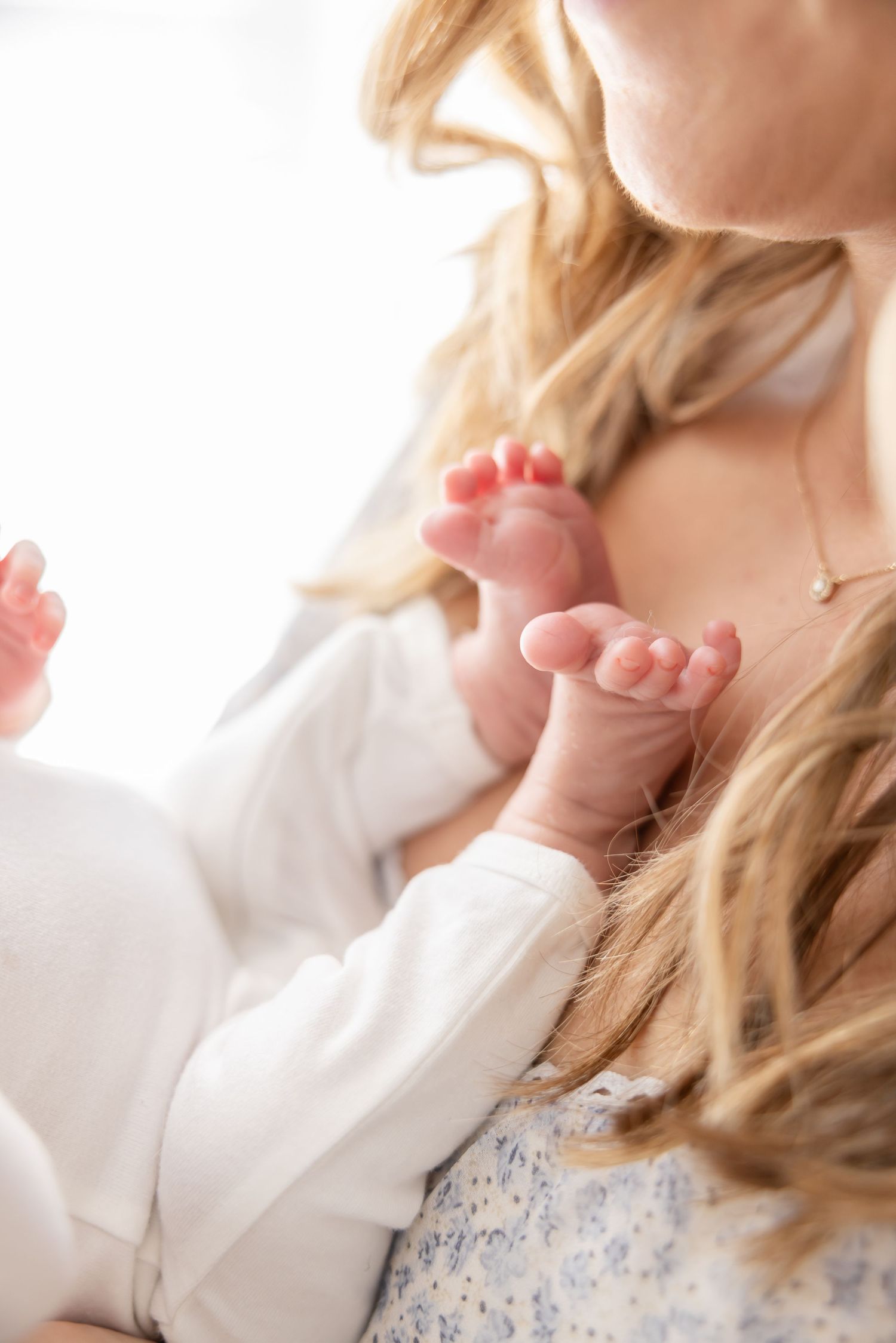 Tiny pink baby feet rest against soft white fabric with blonde hair visible in gentle natural lighting.