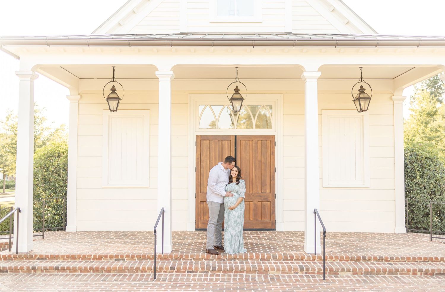 A couple embraces lovingly in front of a classic white chapel with wooden doors and hanging lanterns.