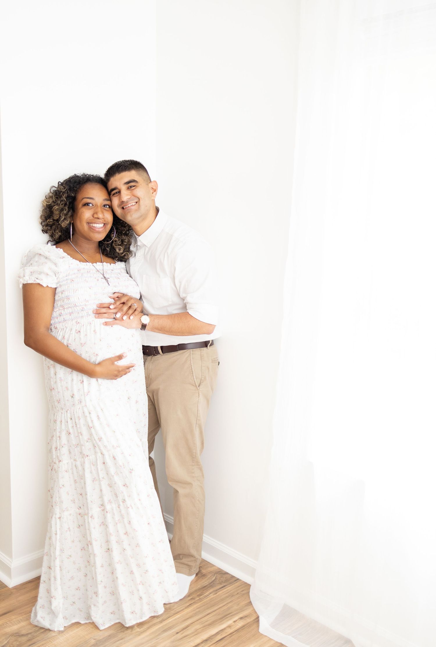 Expectant couple in white dress and khaki pants embrace while standing against a white wall during maternity photoshoot.