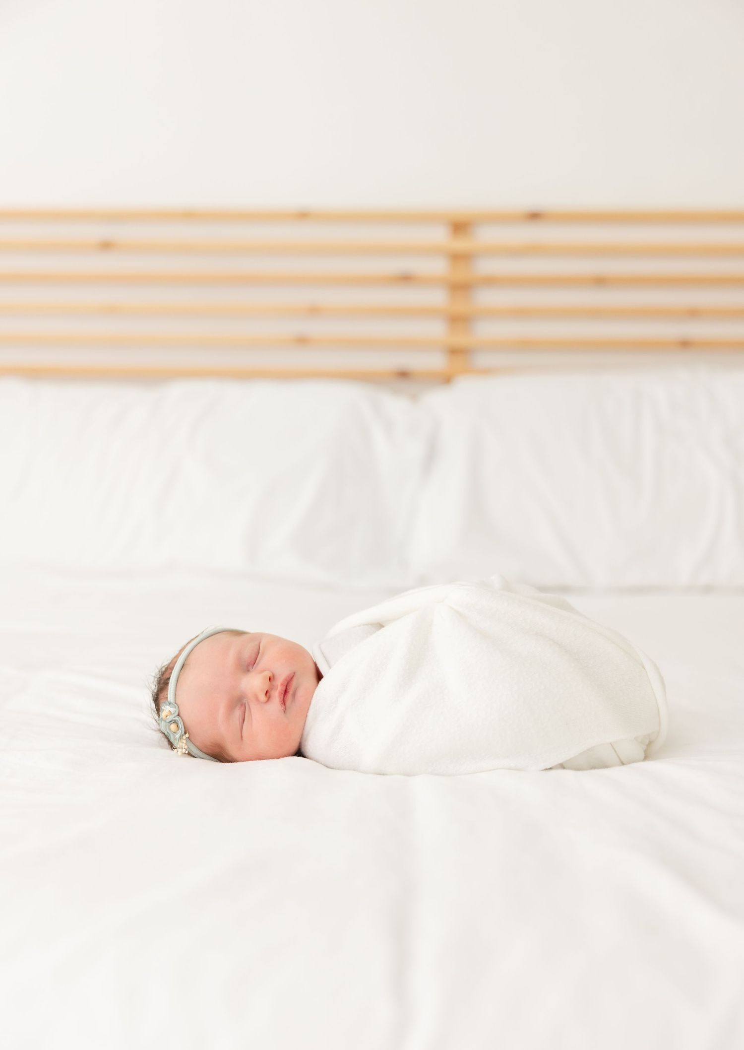 A sleeping newborn baby wrapped in white swaddling cloth lies peacefully on white bedding.
