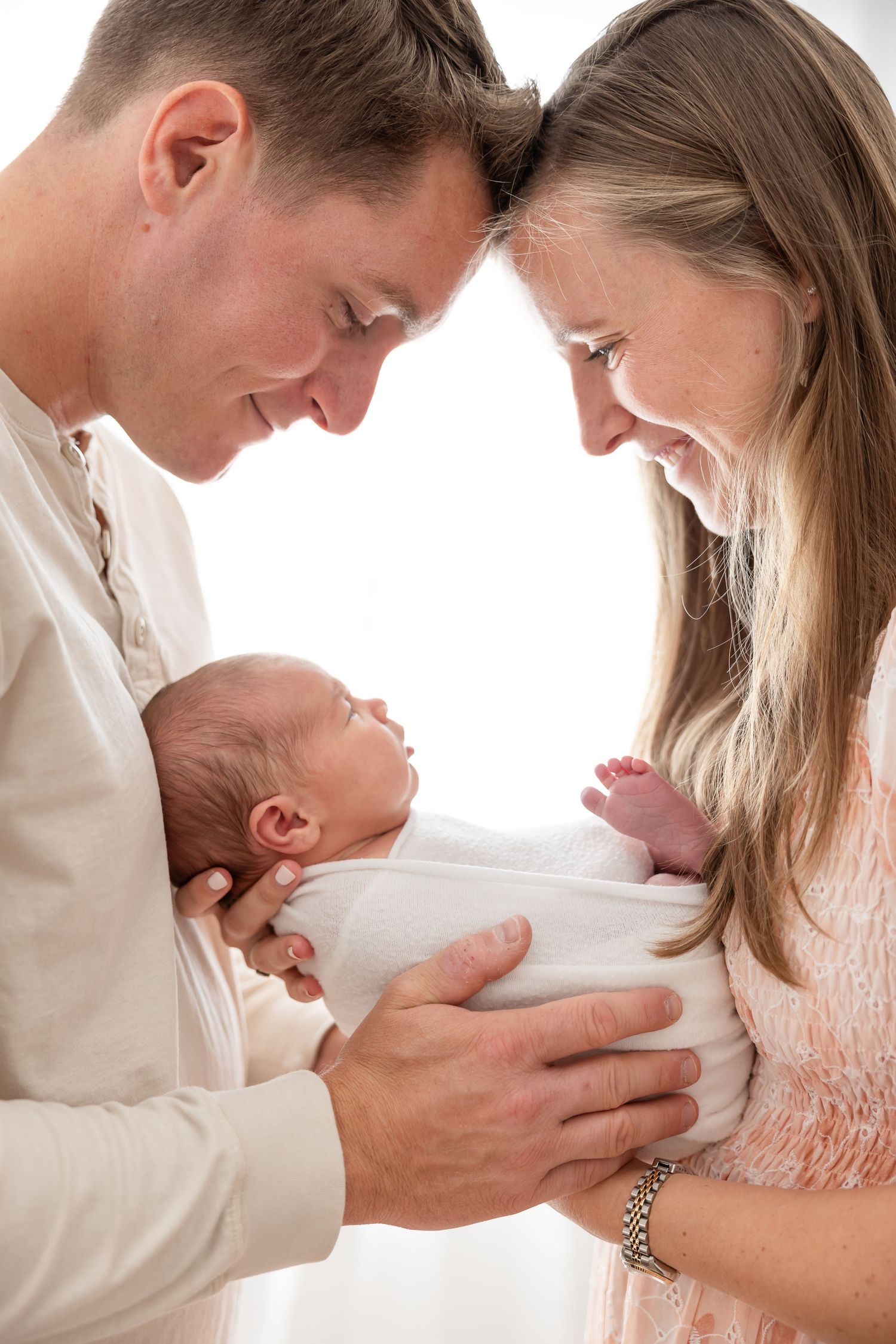 Parents in white clothing lovingly gaze down at their newborn baby wrapped in a blanket against light background.