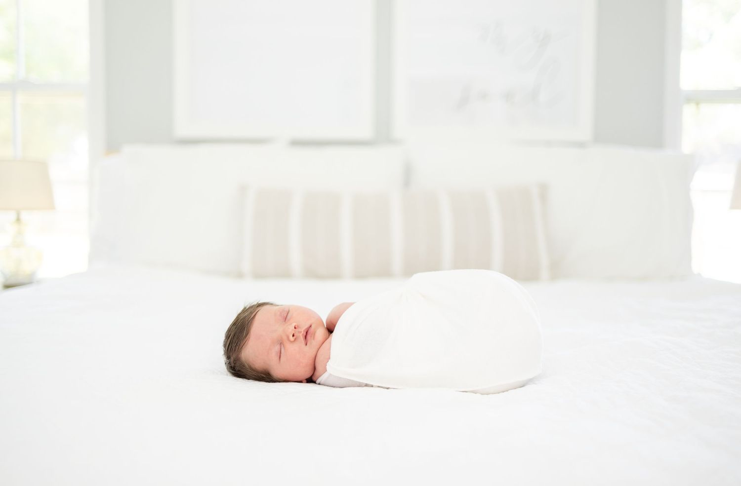 A swaddled newborn baby sleeps peacefully on white bedding in a bright, serene bedroom.