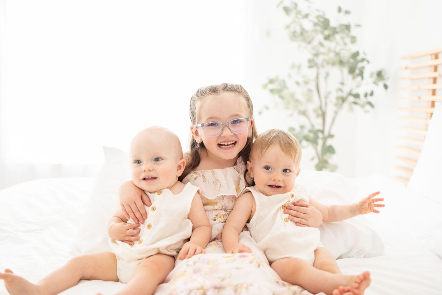 Group of happy babies and toddlers lying in white bed together wearing cream colored outfits.