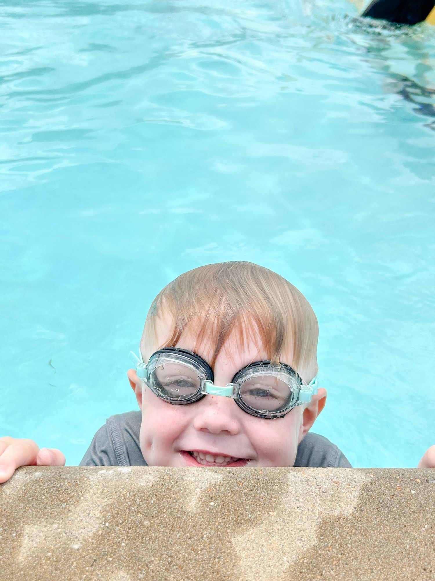 A swimmer wearing gray goggles peeks up at the edge of a bright blue swimming pool while smiling.