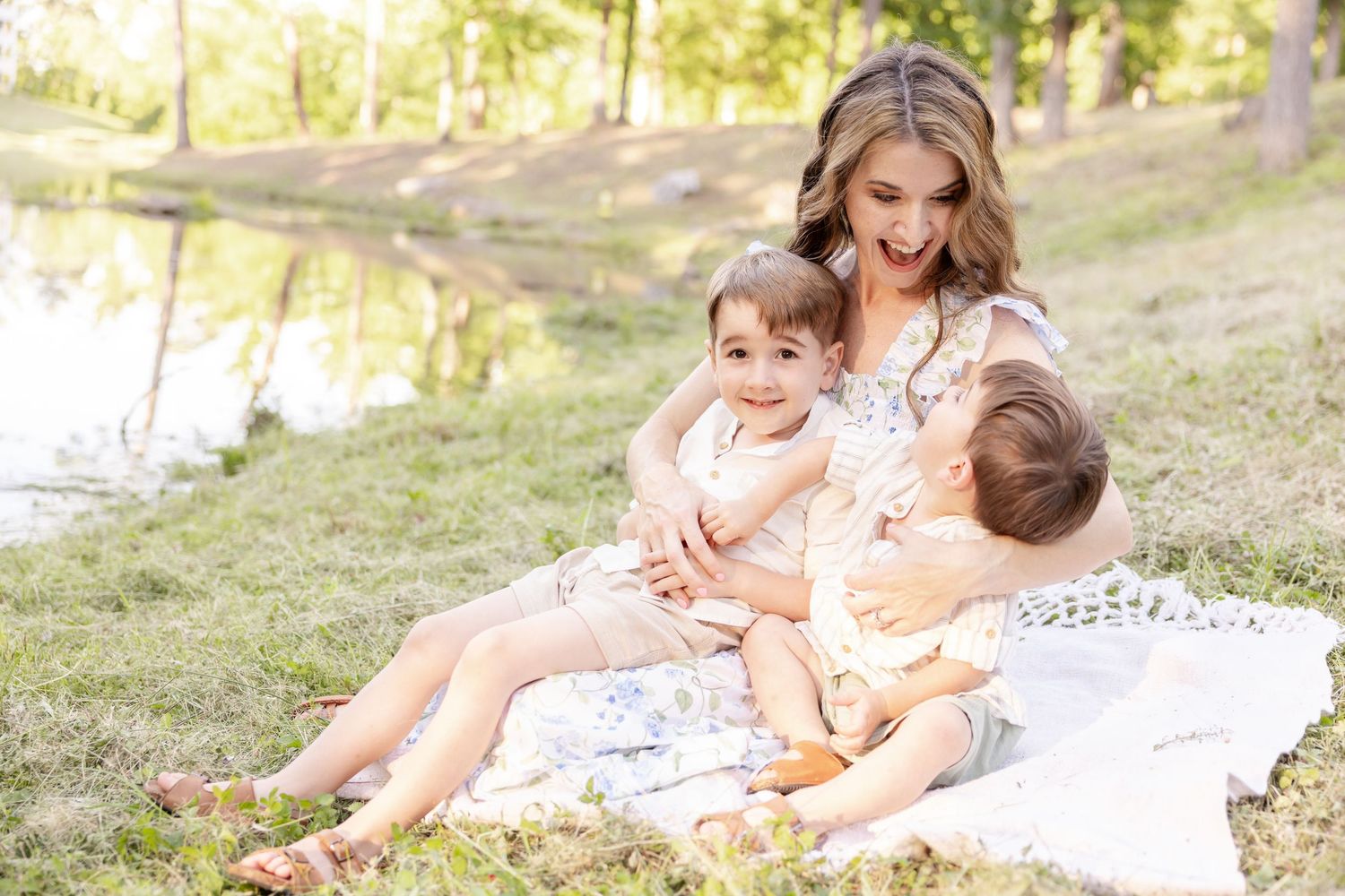 A loving scene by a pond with people sitting together on a white blanket outdoors on a sunny day.