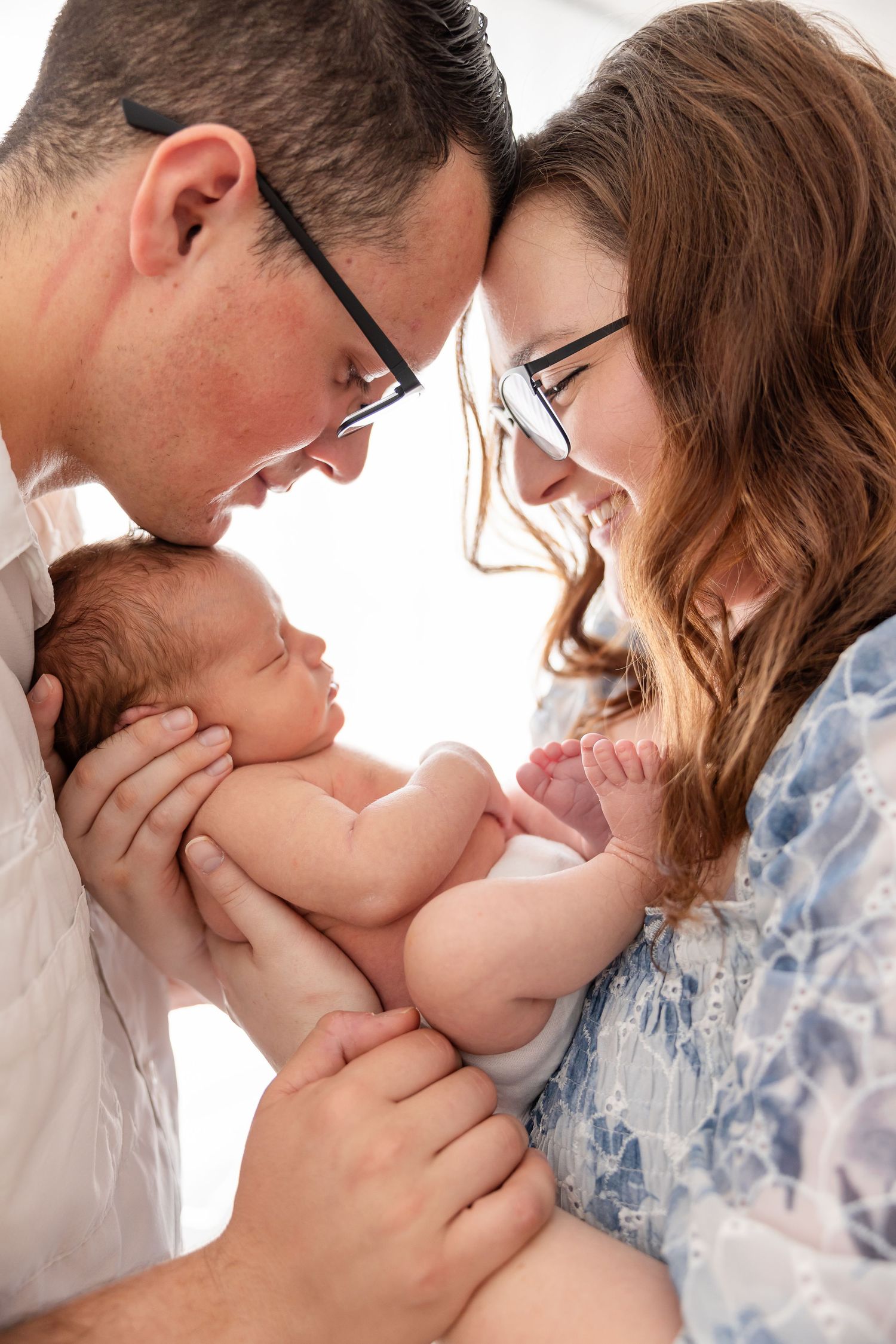 Parents in glasses lovingly gaze down at their sleeping newborn baby in tender intimate family portrait.