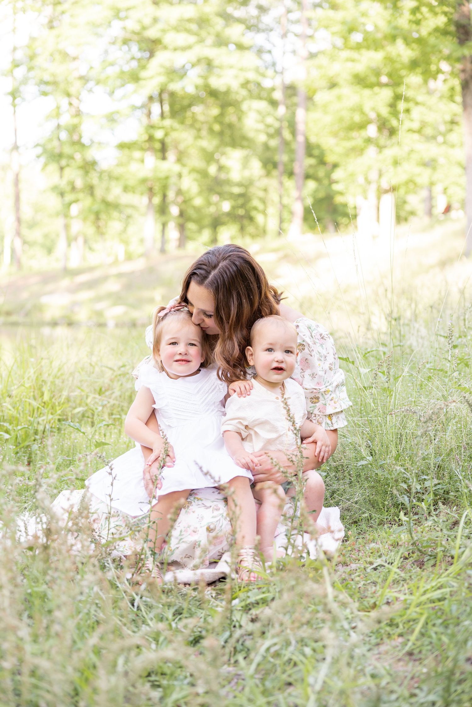 A tender outdoor family moment captured in a sunlit meadow with soft wildflowers and greenery in the background.