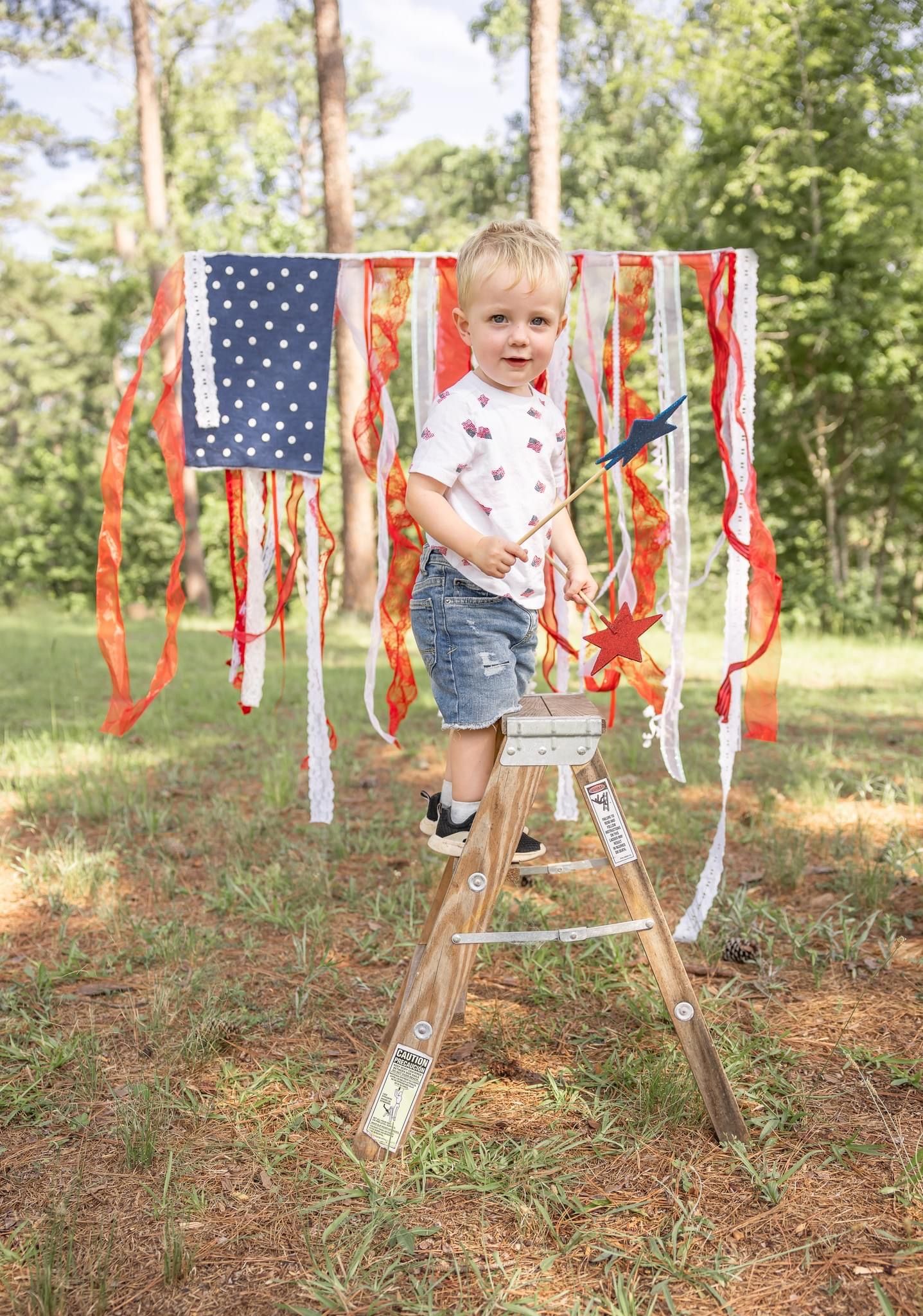A small child stands on a wooden stepladder in front of decorative American flags and red ribbons hanging in a wooded setting.