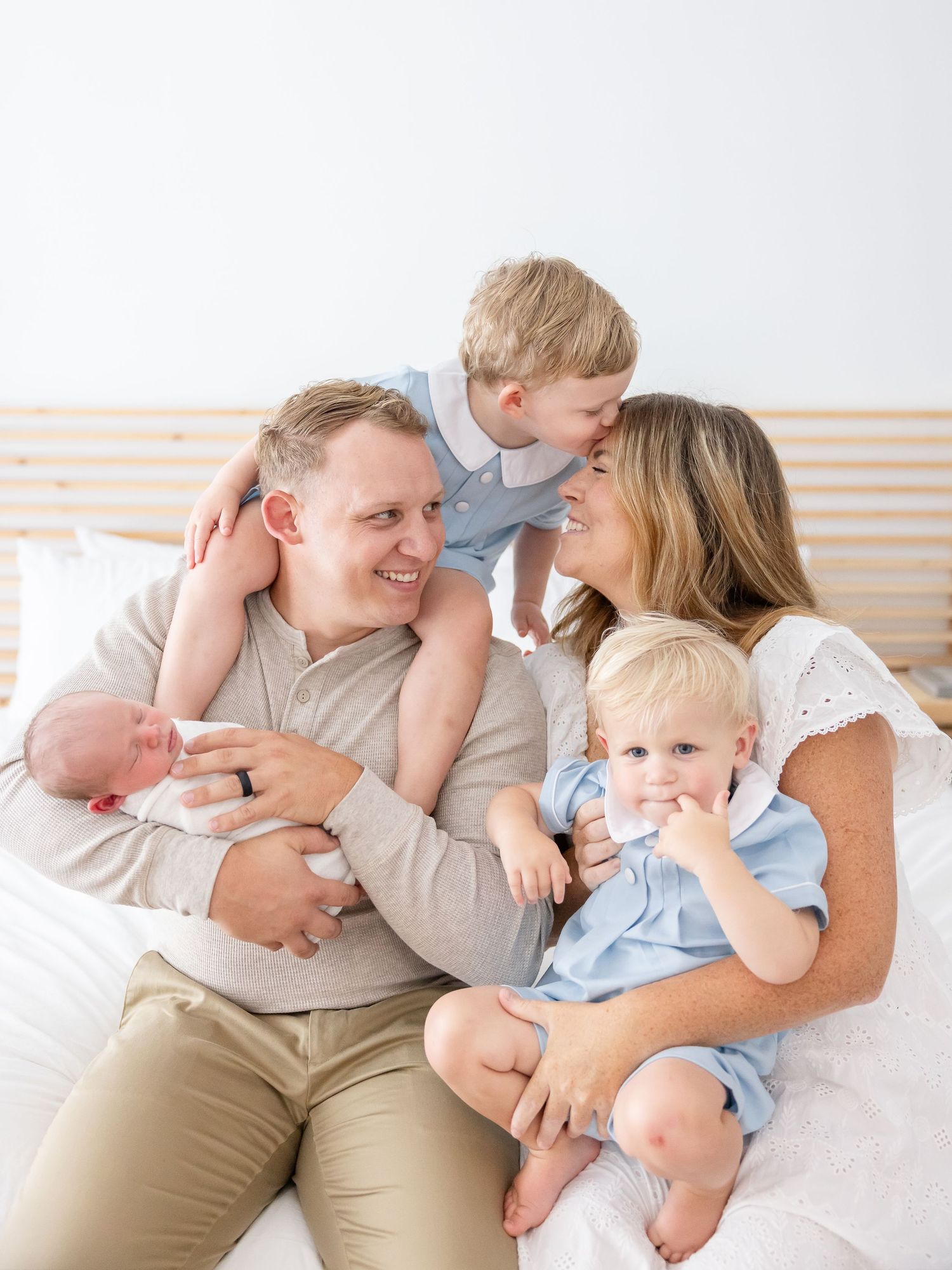 A family of five sits together on a white bed, hugging and laughing in a light and bright room at their Columbus GA newborn portrait session