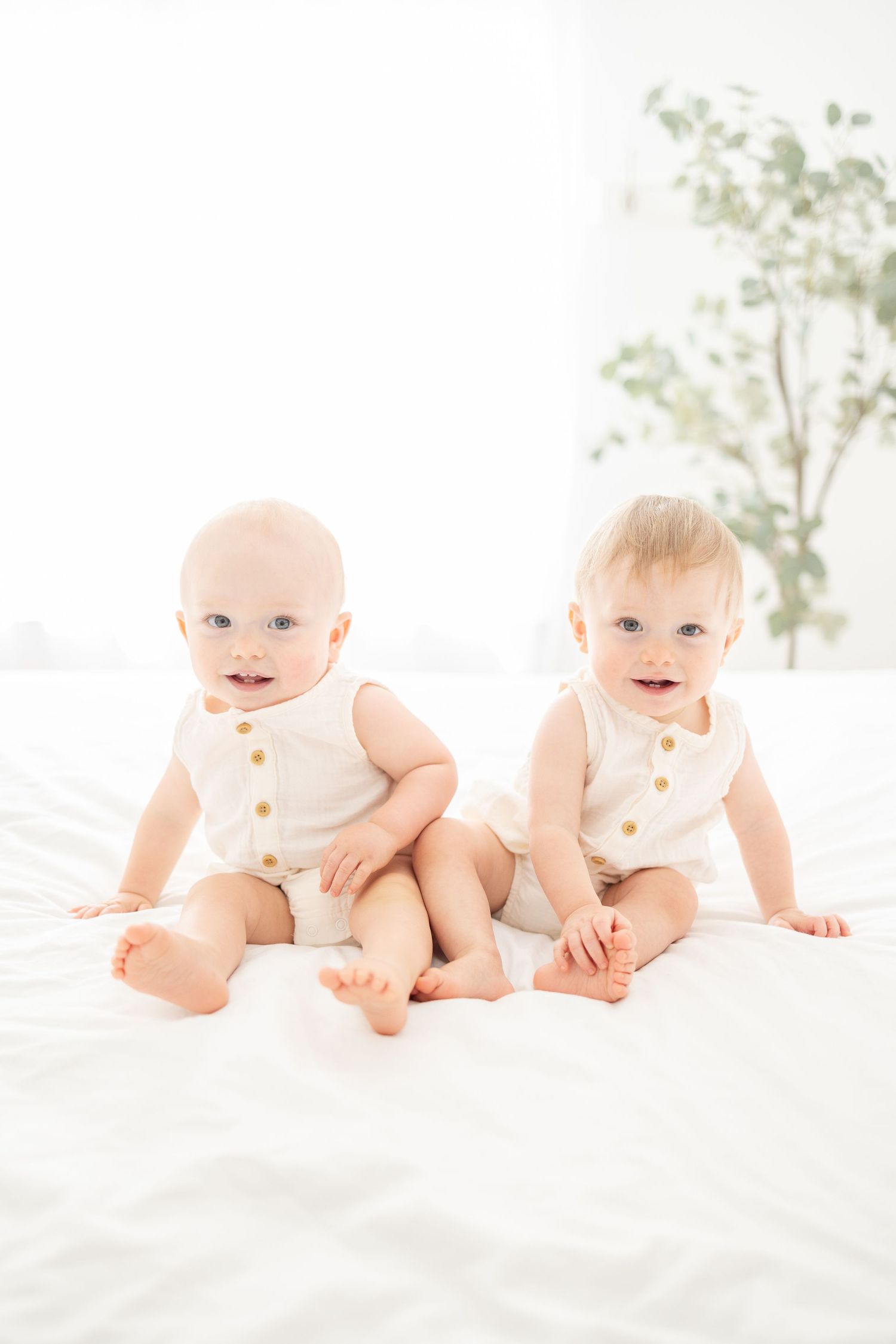 Two adorable infants in white button-up rompers sit together on white bedding with greenery in background.