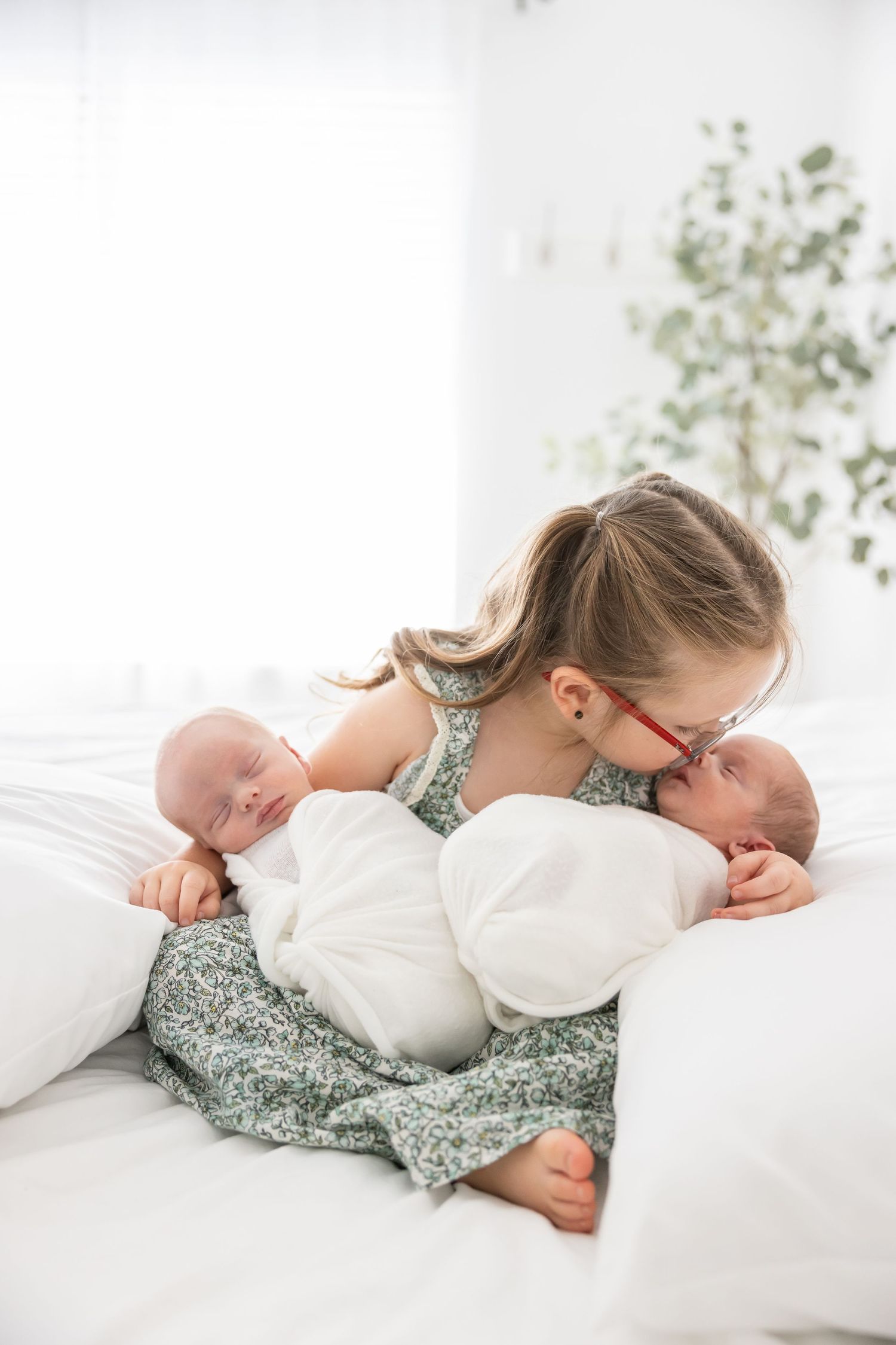A young person in a floral dress lies on white bedding with two swaddled newborn babies.