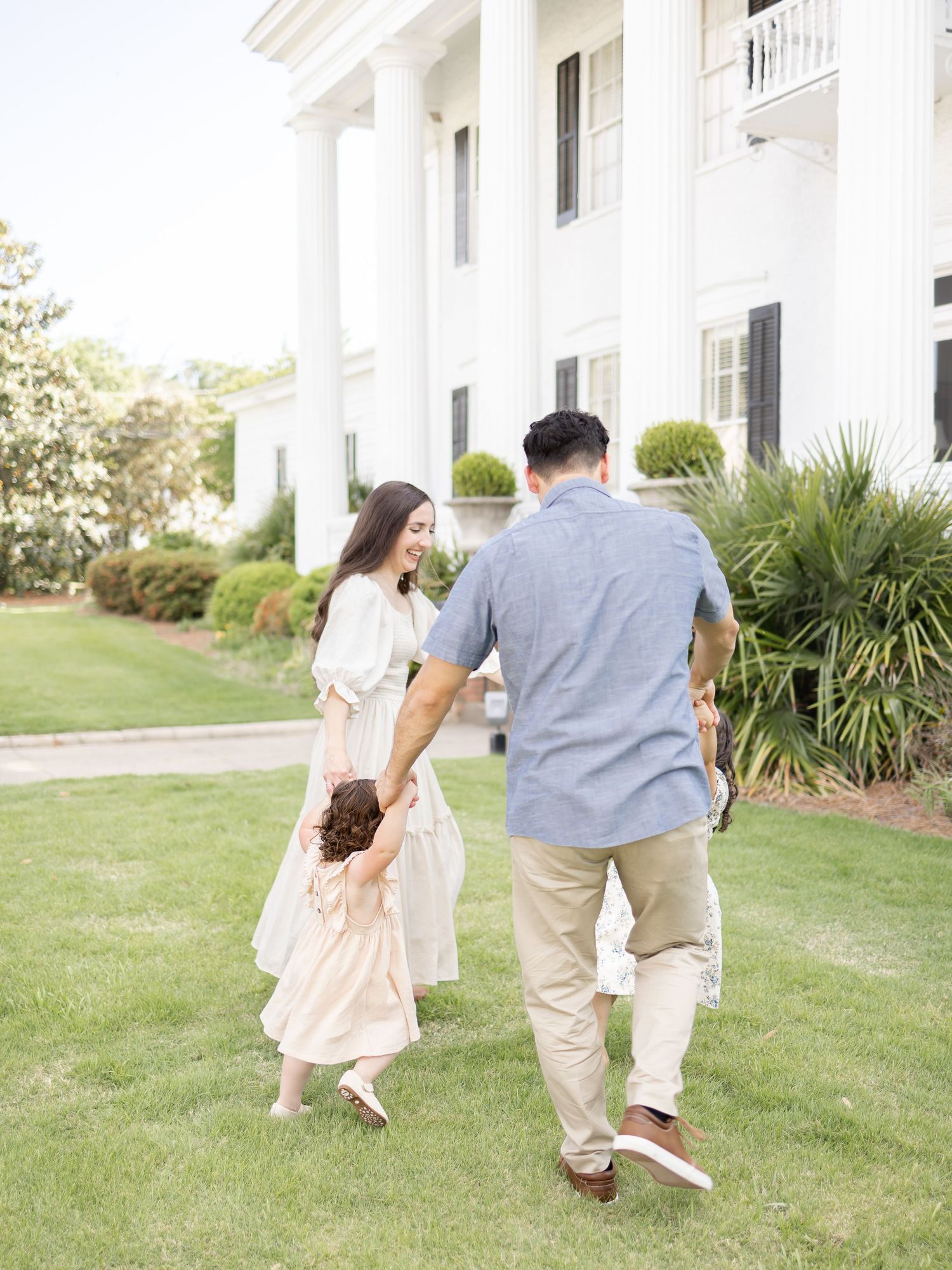 Family walking together on a green lawn in front of a white colonial-style mansion with white columns.