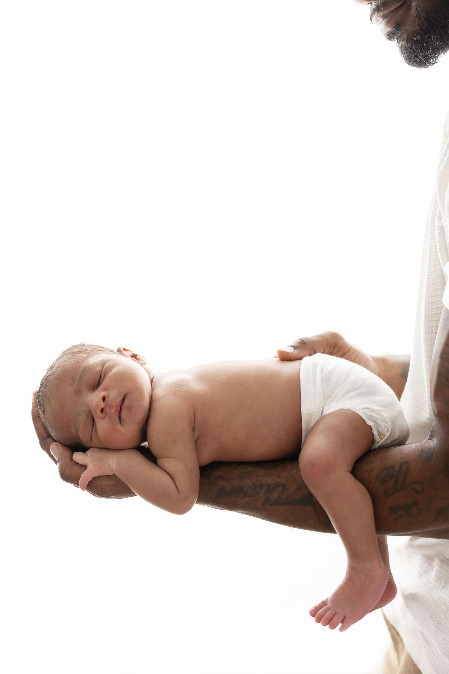 A sleeping newborn baby wearing a diaper rests peacefully on a tattooed forearm against a white background.