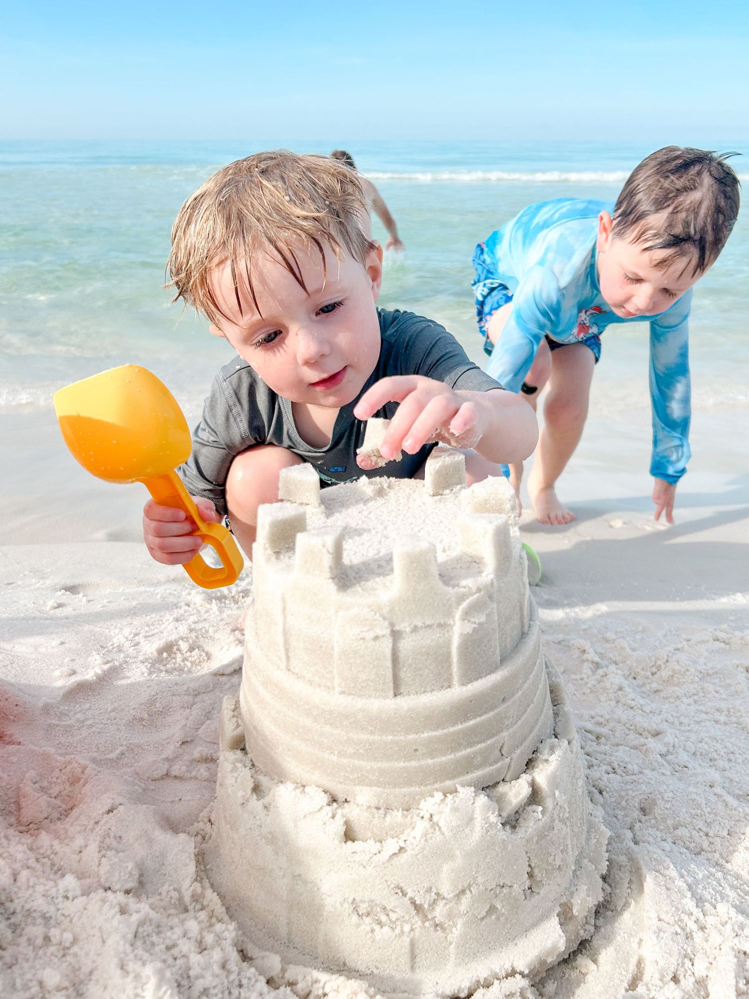 Two children playing on a sunny beach with sand toys and building a sandcastle near the ocean waves.