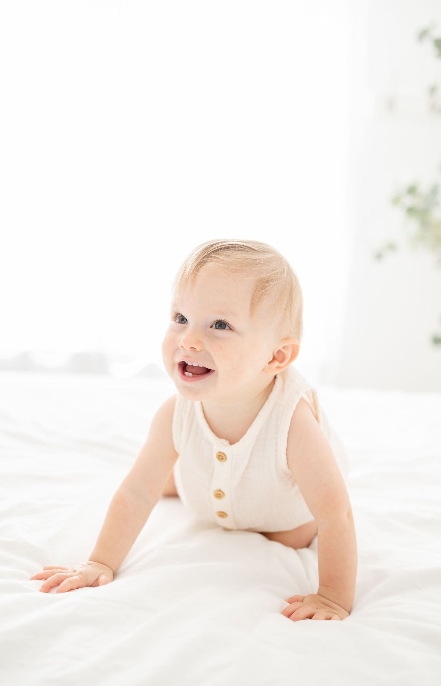 Baby crawling on white bedding wearing sleeveless white onesie while smiling and playing.