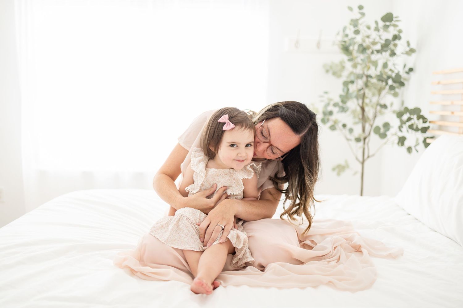 A loving parent and child share a tender moment together while sitting on a white bed with a plant in the background.