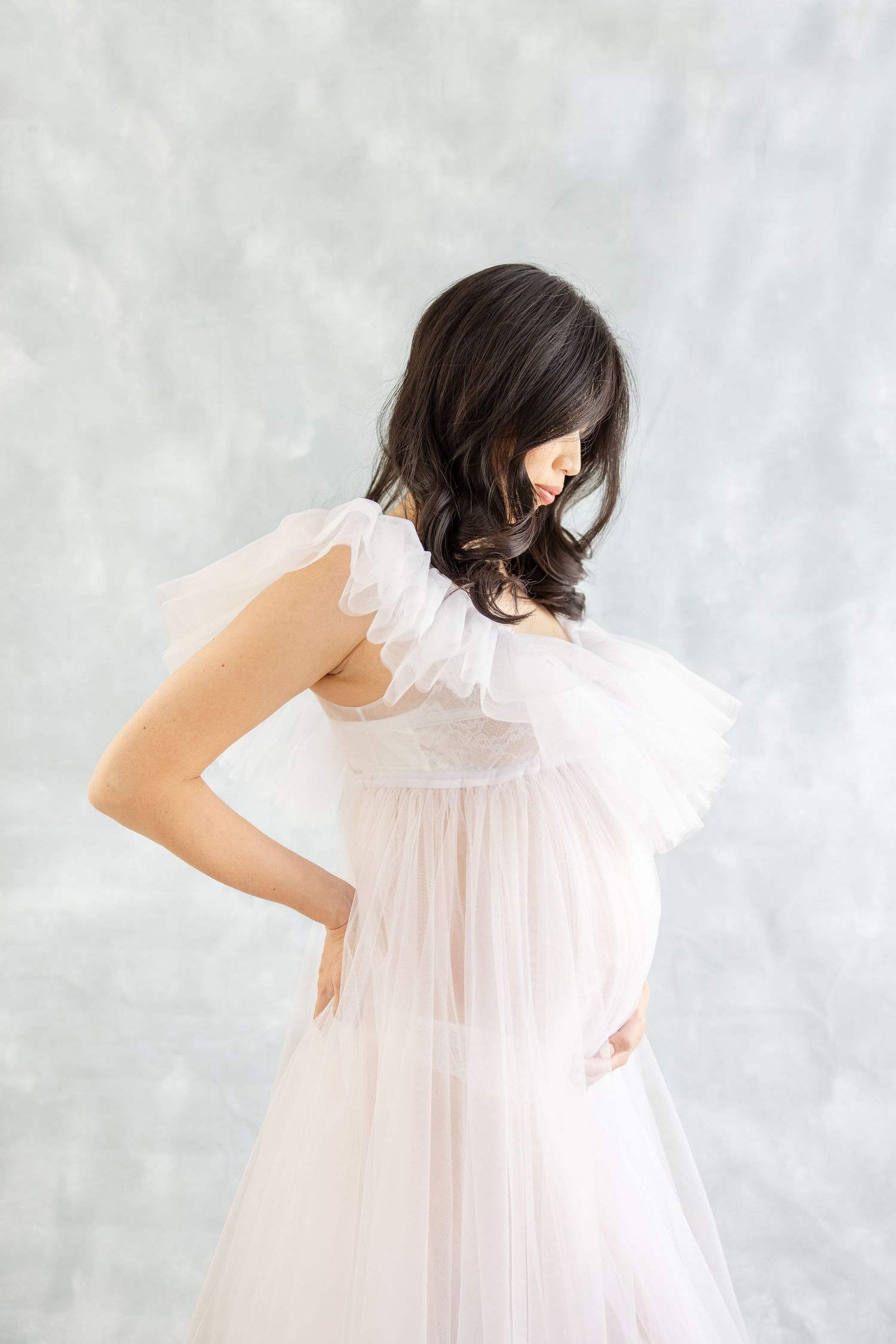 Ethereal white tulle maternity dress with ruffled neckline captured in dreamy soft light against neutral backdrop.