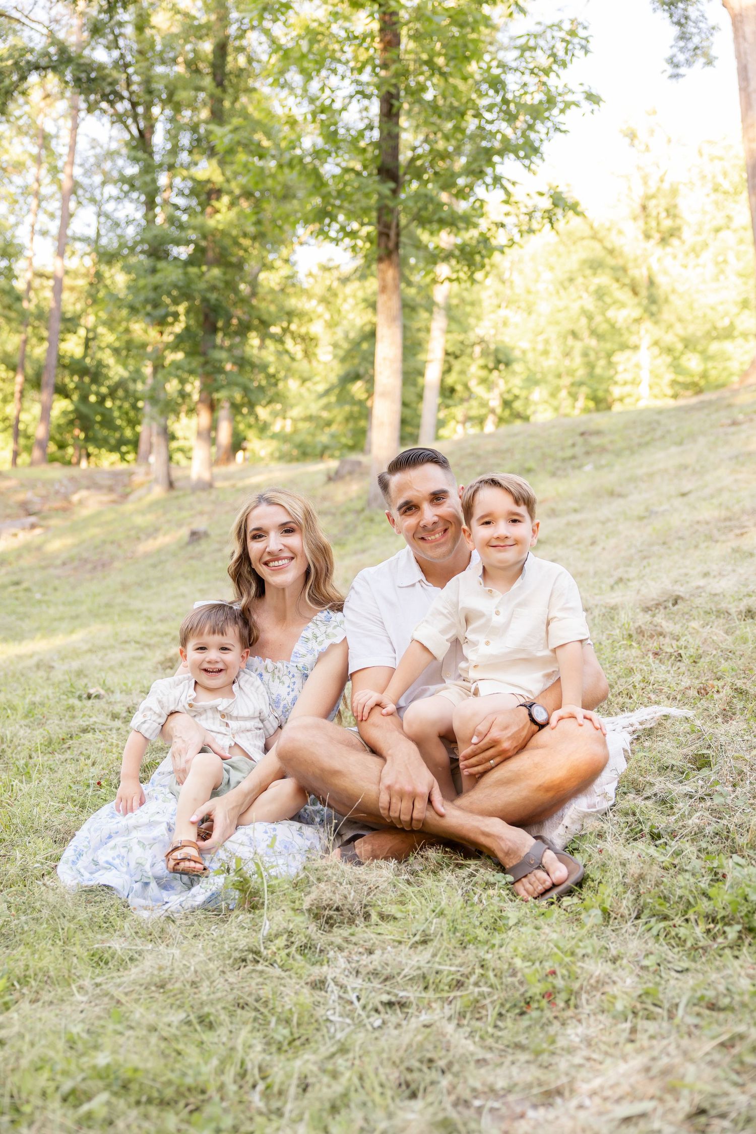 Family relaxes together on grassy hillside during golden hour photoshoot in wooded setting.