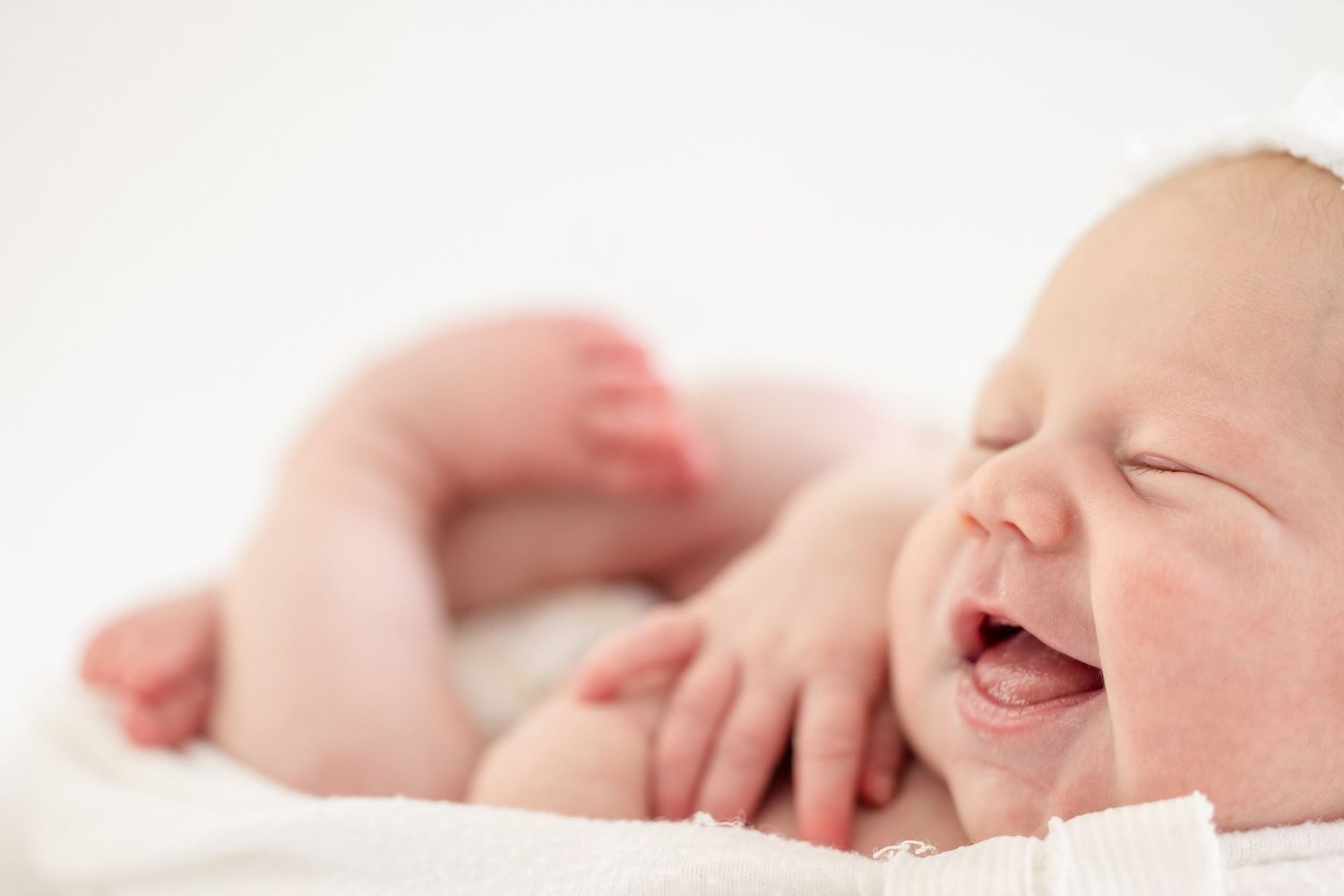 A joyful baby lies on white bedding with closed eyes and a big laugh, showing pure happiness.