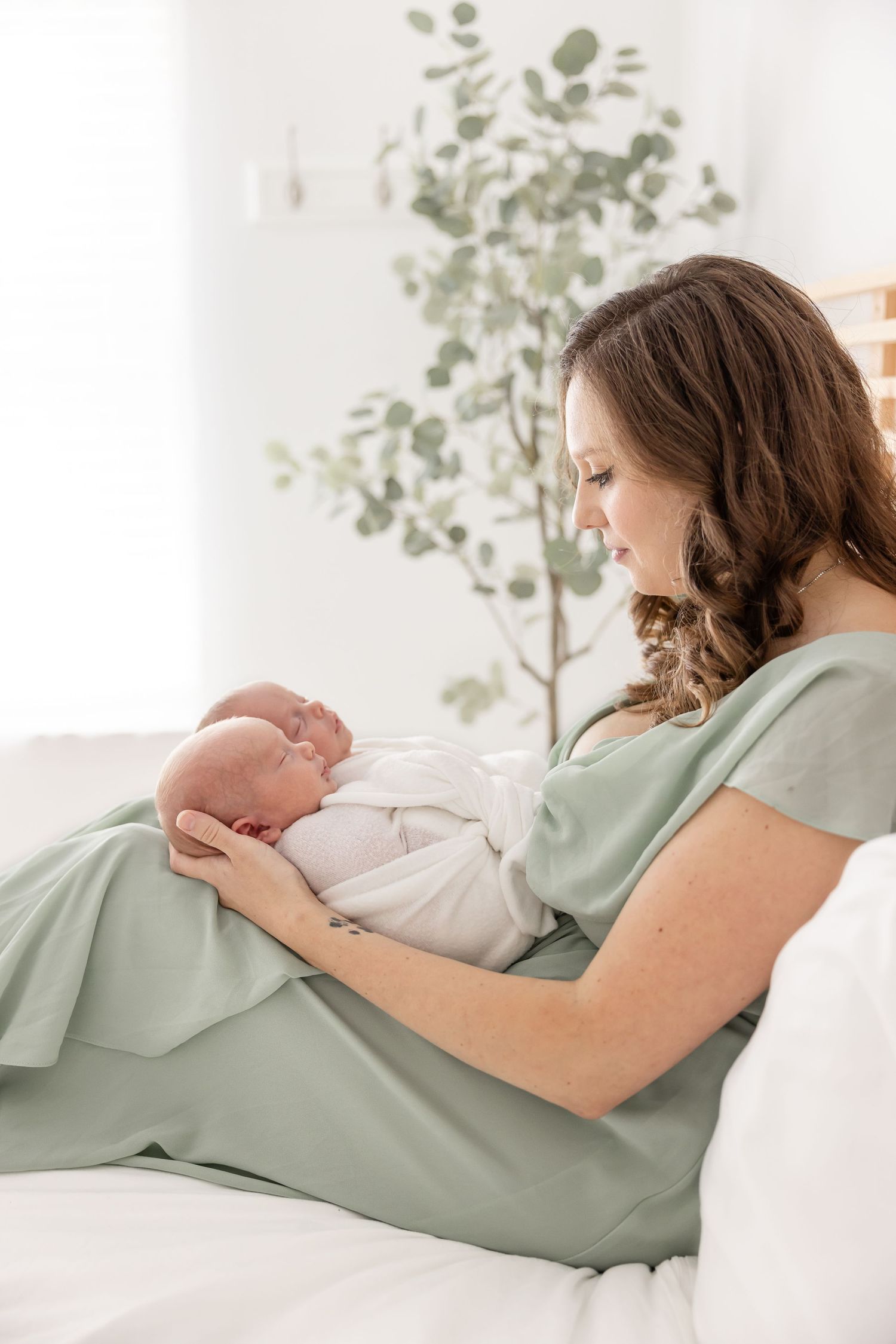 Woman in sage green dress sits on white couch holding newborn baby in peaceful nursery setting with eucalyptus plant.