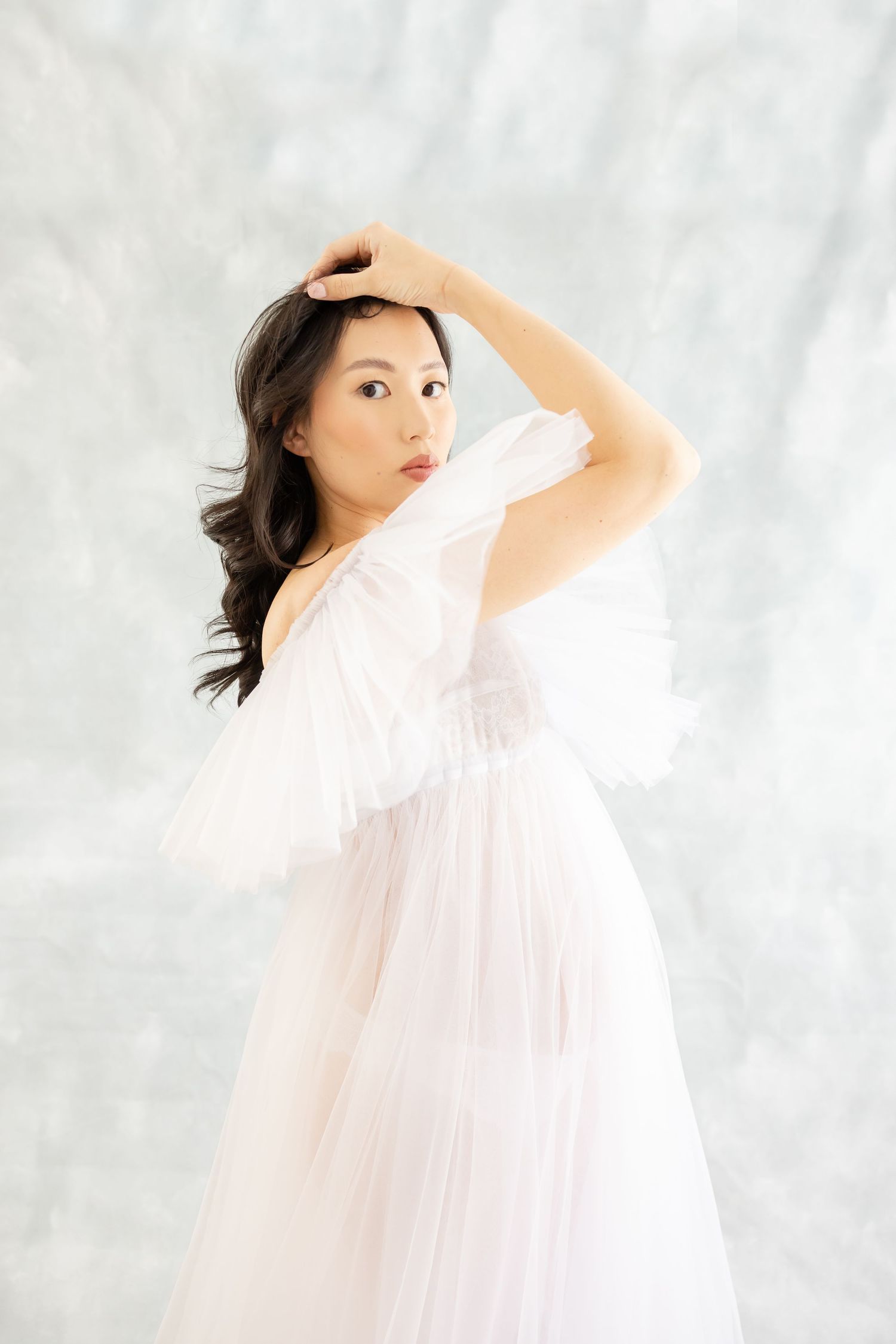 A person in a flowing white dress poses gracefully against a light gray backdrop in an ethereal portrait.