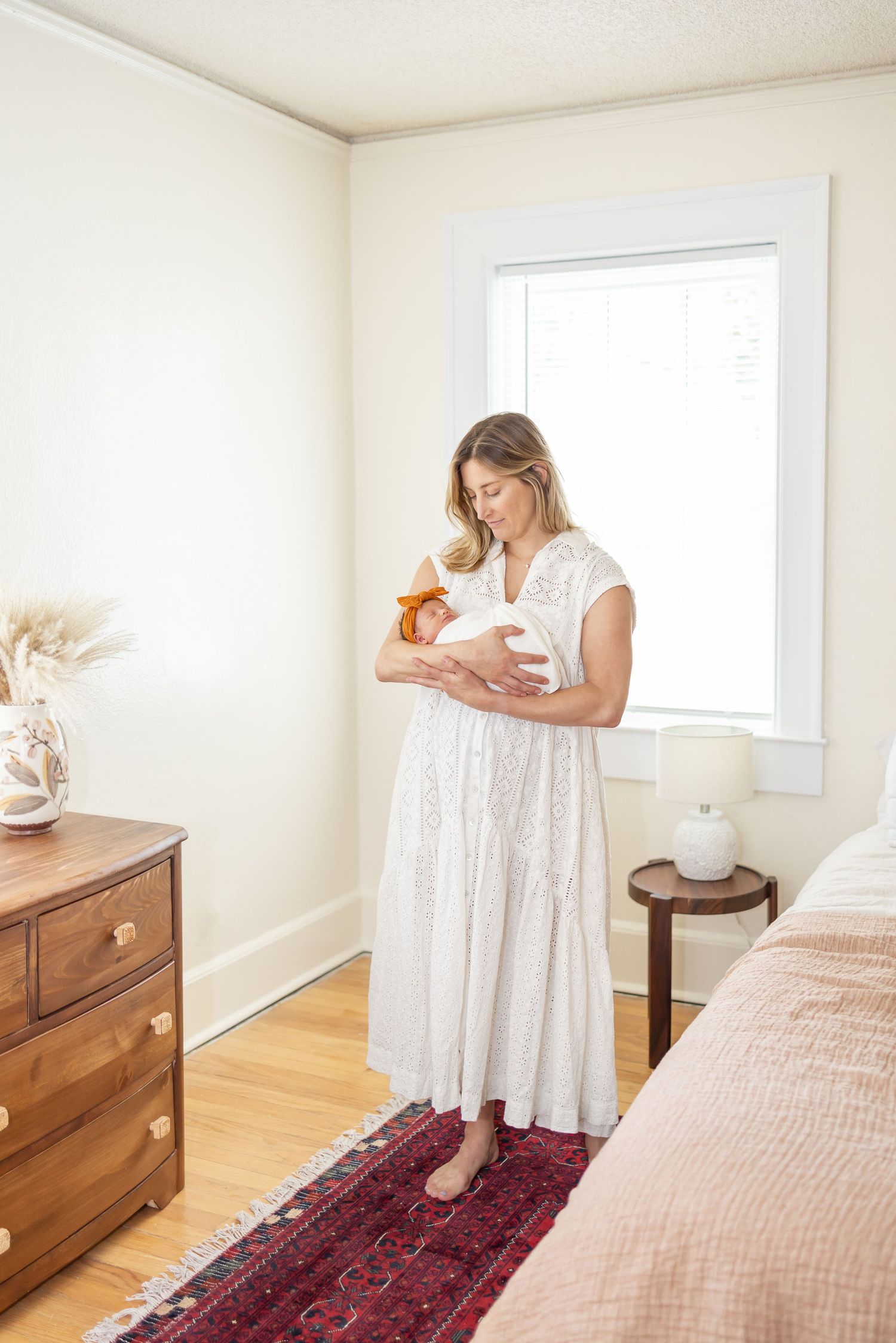 A person in a white dress stands by a window in a bedroom with wood floors and a vintage runner rug.