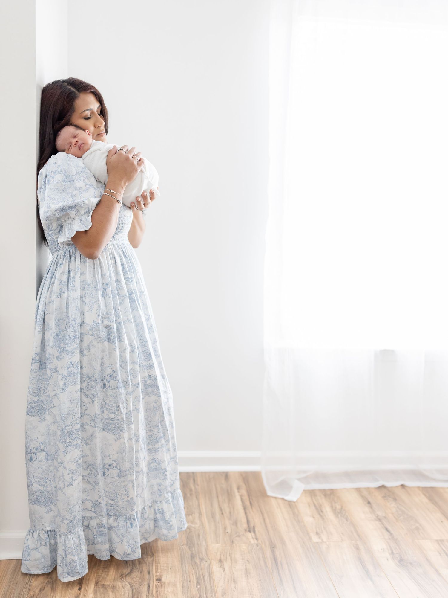 Woman in a flowy blue and white floral print dress stands holding a small newborn in a bright airy room.