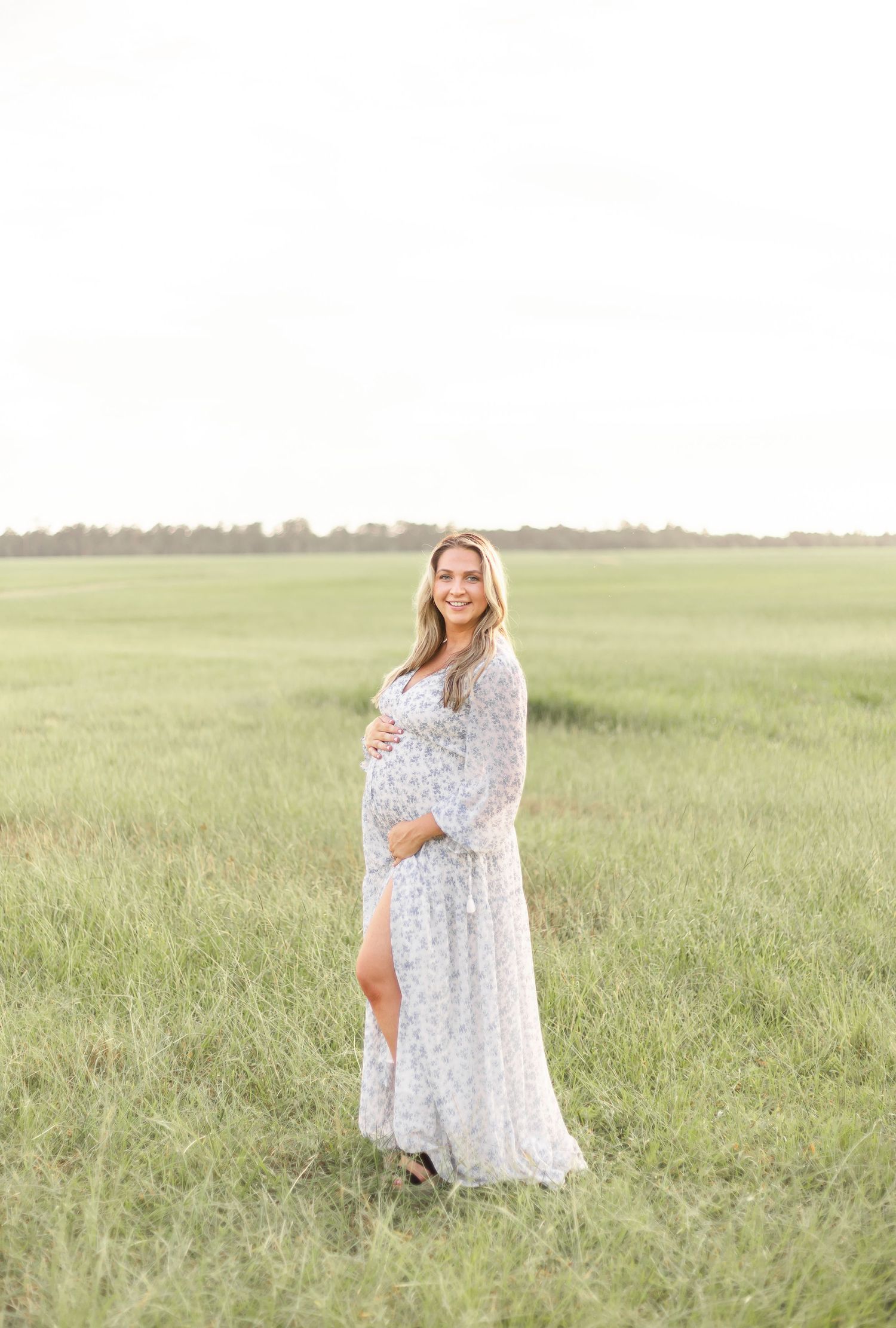 A person in a flowing white floral dress stands in a sunlit meadow of tall grass during golden hour.