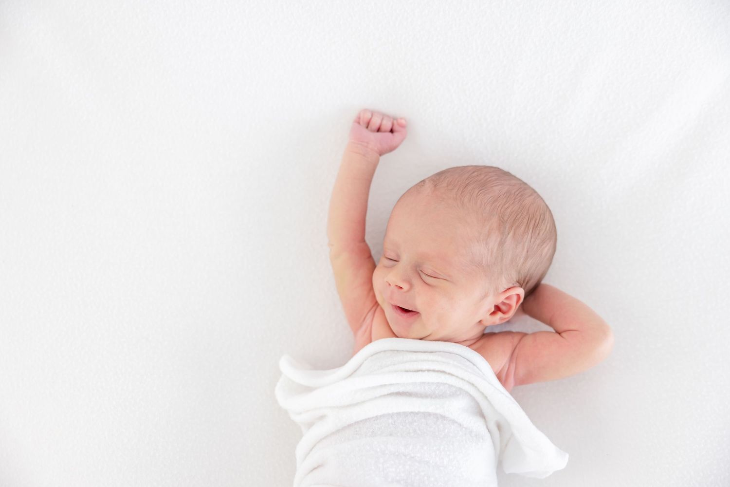 Sleeping newborn baby stretches arm upward while wrapped in white blanket on light background.
