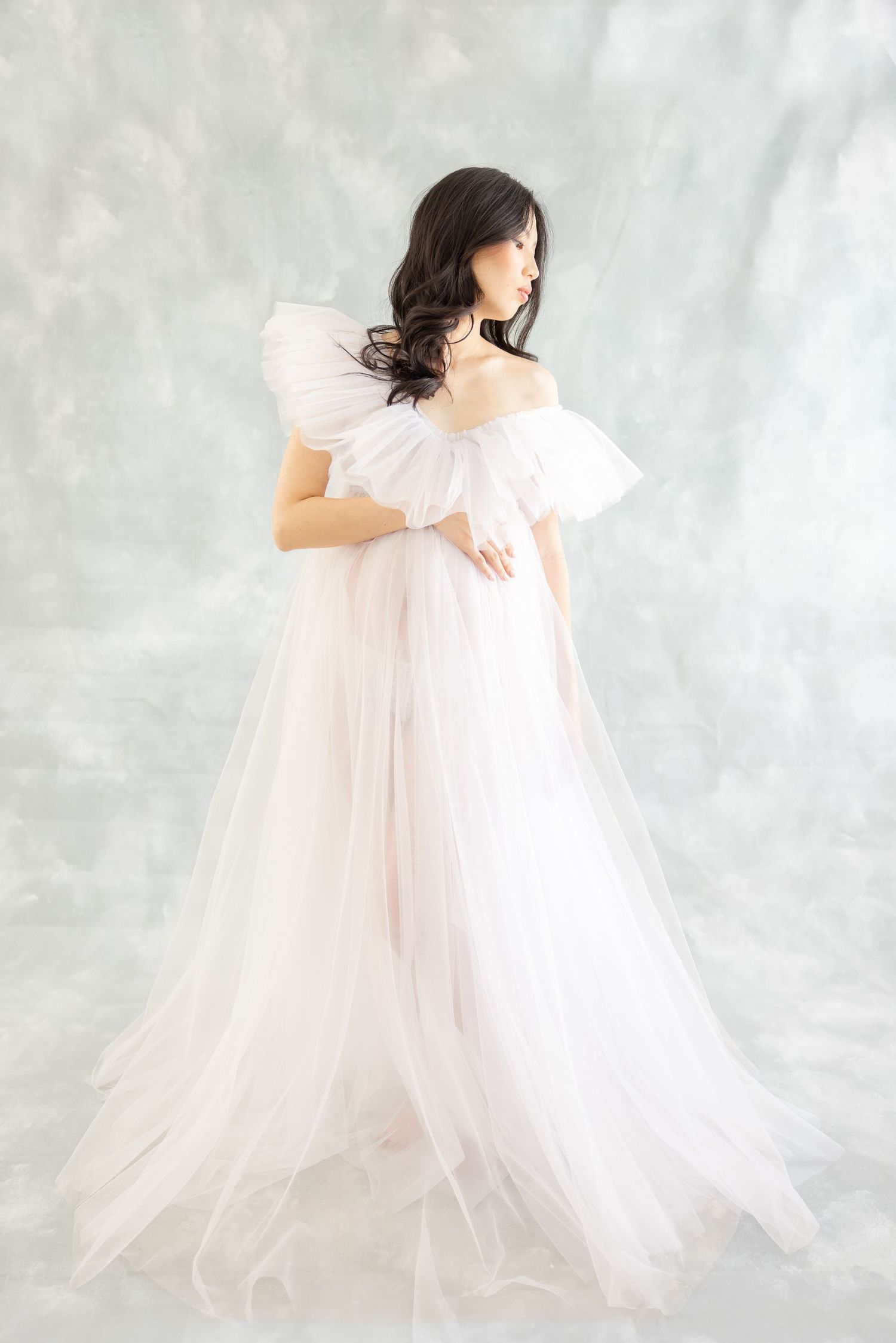 A person in a romantic off-shoulder blush pink tulle gown poses elegantly against a light gray backdrop.