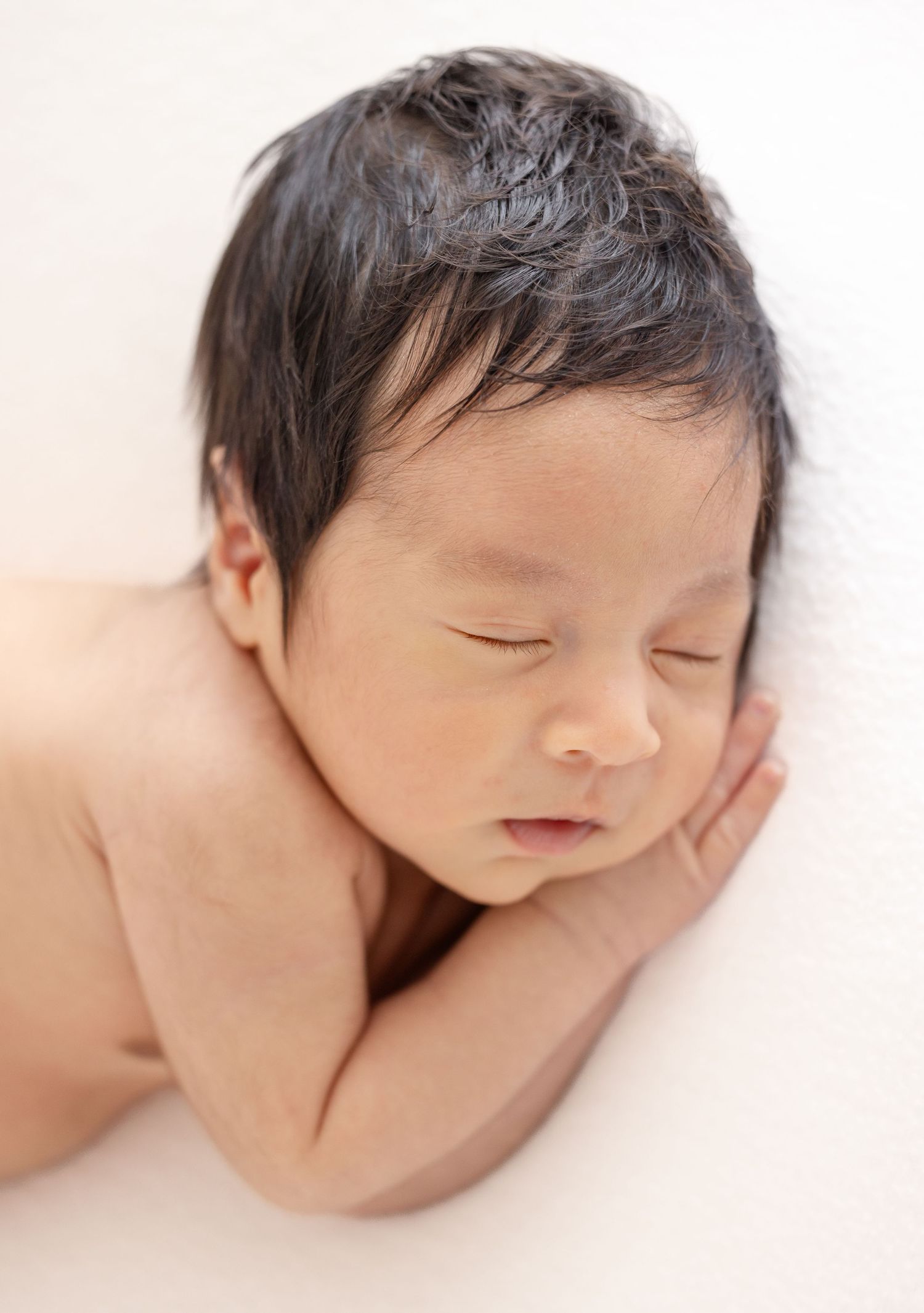 Peaceful sleeping newborn baby with dark hair rests on soft white blanket during professional photography session.