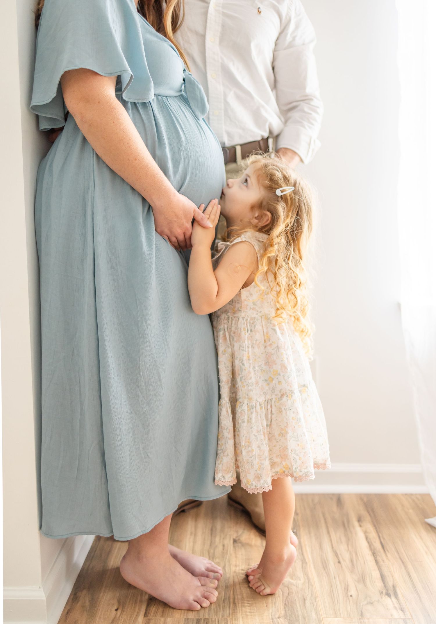 Young child in floral dress kisses expecting mother's belly while standing, wearing light blue maternity dress indoors.