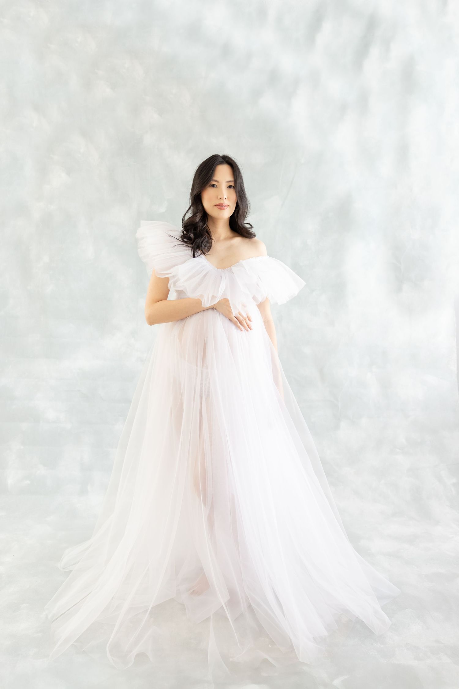 A person in an ethereal white tulle gown poses elegantly against a light gray backdrop.