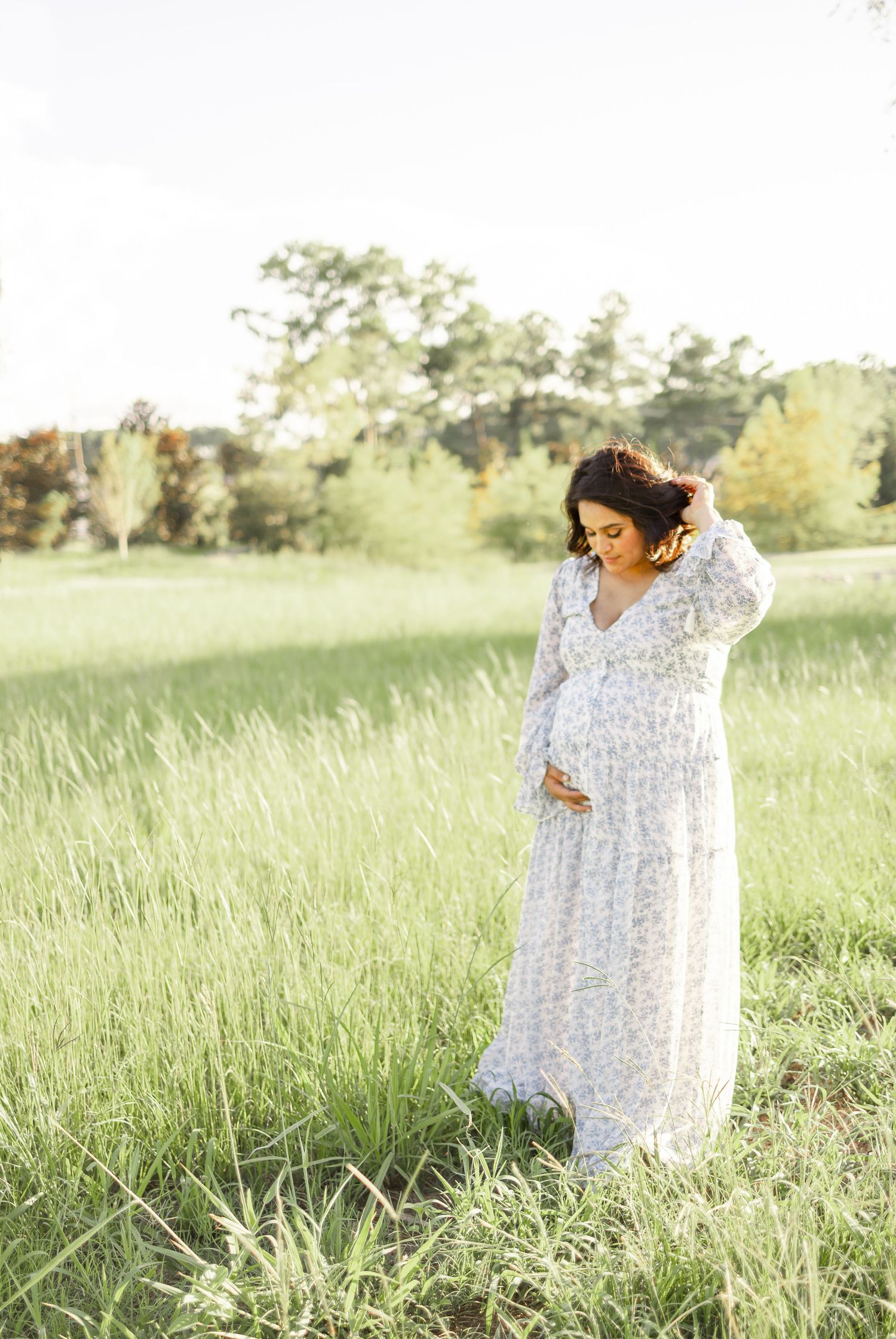 A pregnant woman in a flowing white floral maxi dress stands in a sunlit field of tall grass during golden hour.