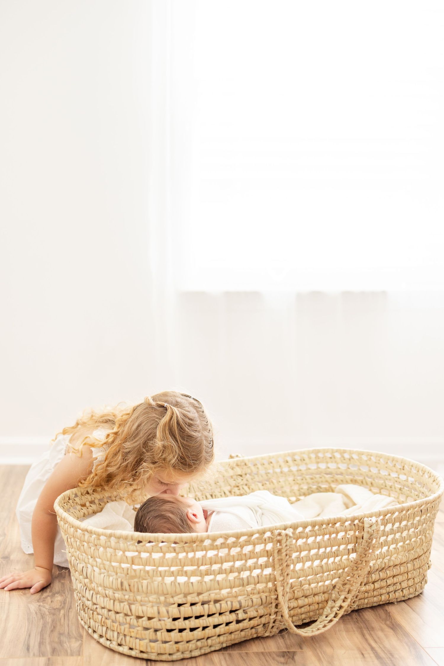 A woven basket bassinet sits on wooden flooring while a child peers inside at a resting baby.