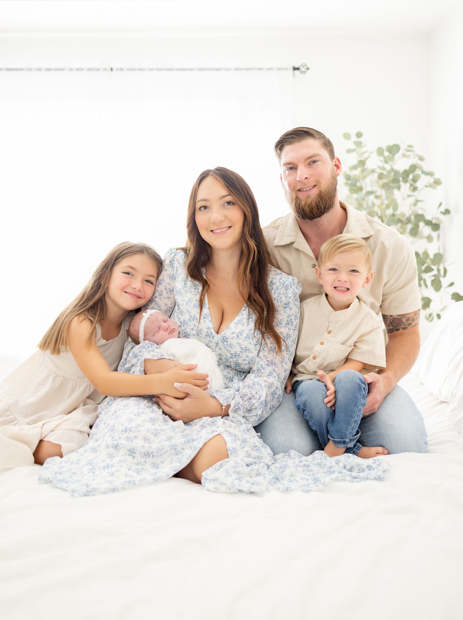Family wearing white and blue sits together on a bed in a bright minimalist room with plants in background.
