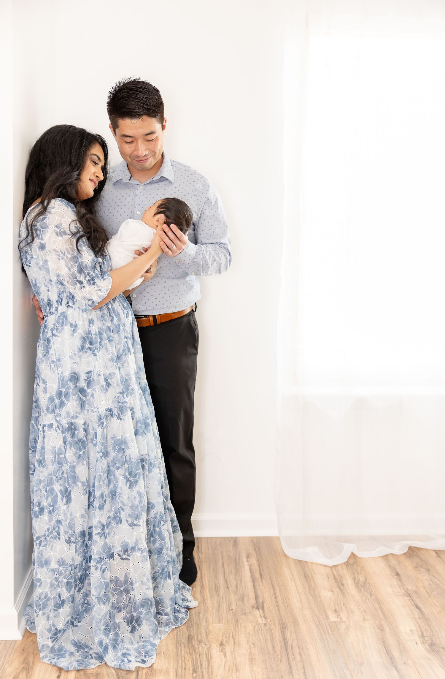 A couple in blue and grey attire stands against a white wall while looking down at something together.