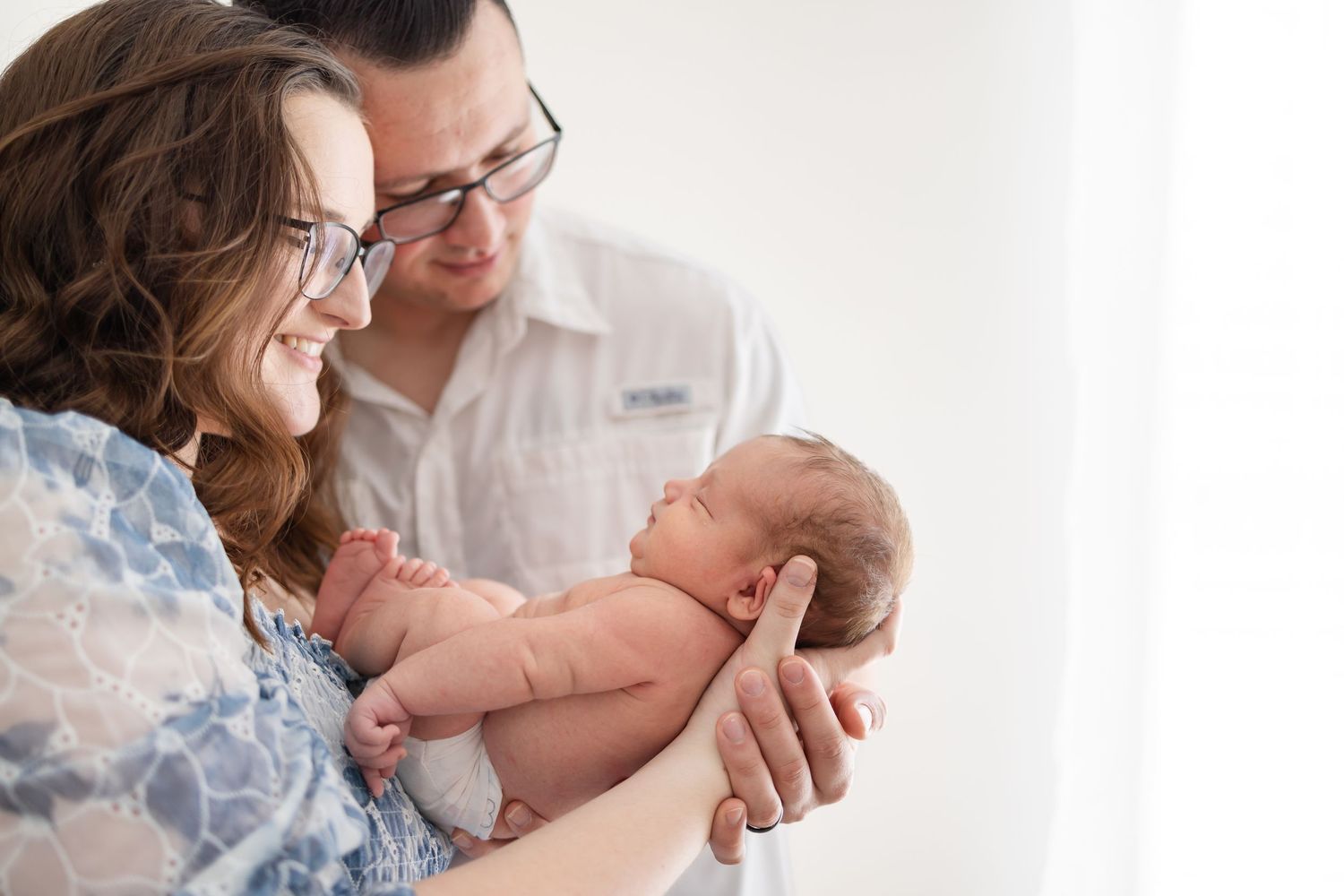 Happy parents hold and look lovingly at their newborn baby against a white wall.