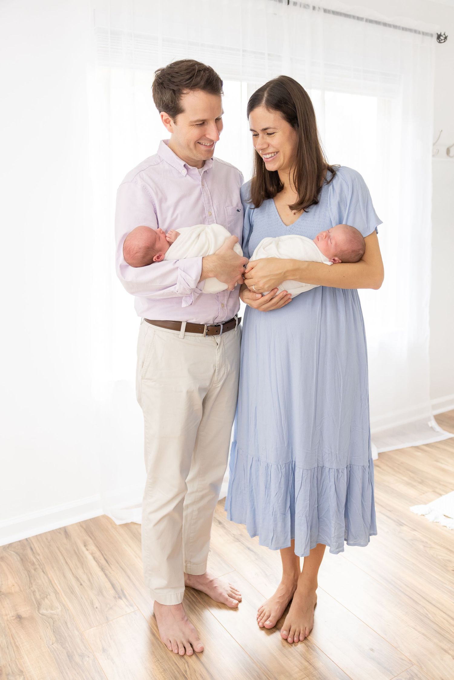 Two parents standing barefoot on hardwood floor hold newborn twins wrapped in white blankets.