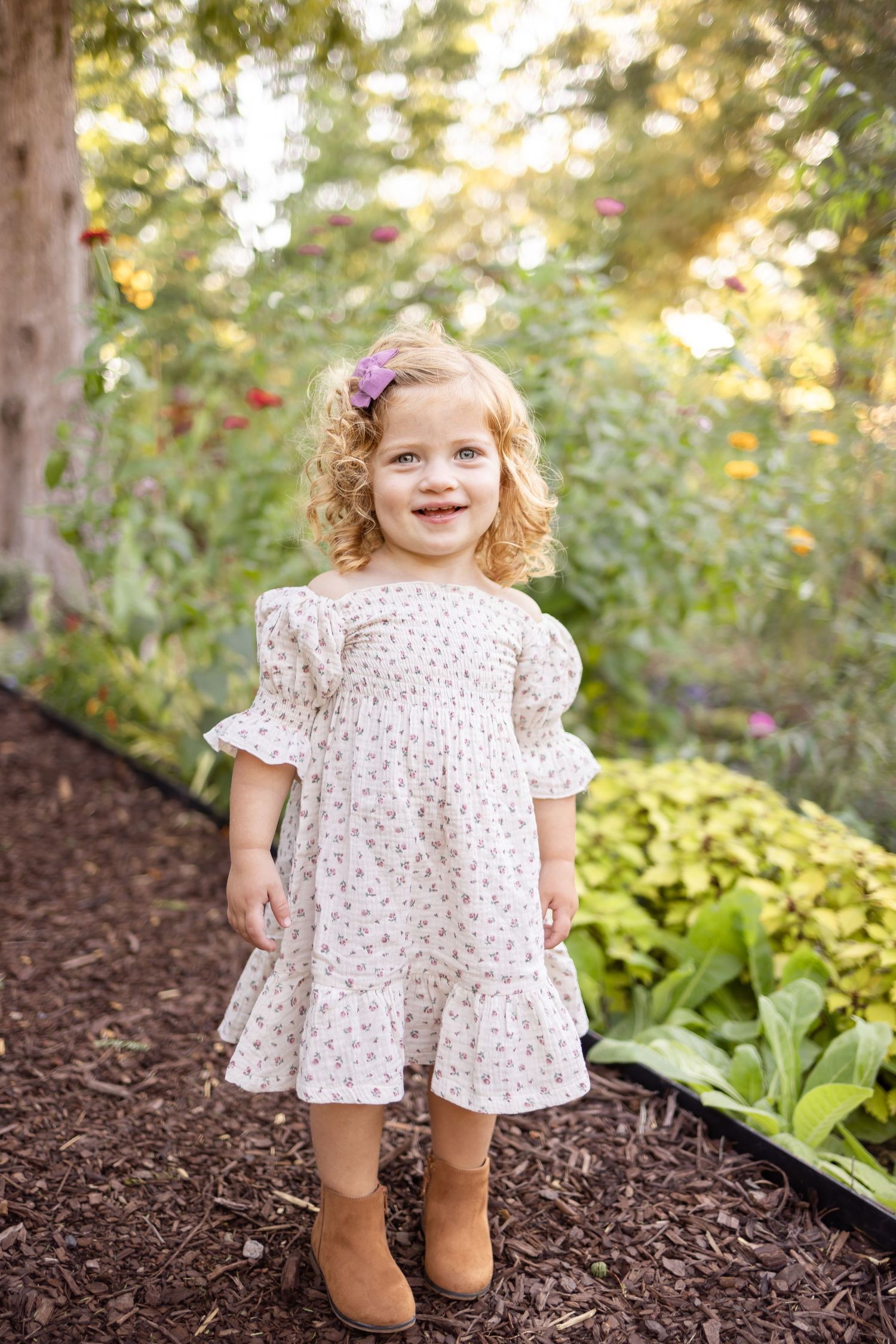 Young child in floral dress and brown boots stands on garden path surrounded by greenery and dappled sunlight.