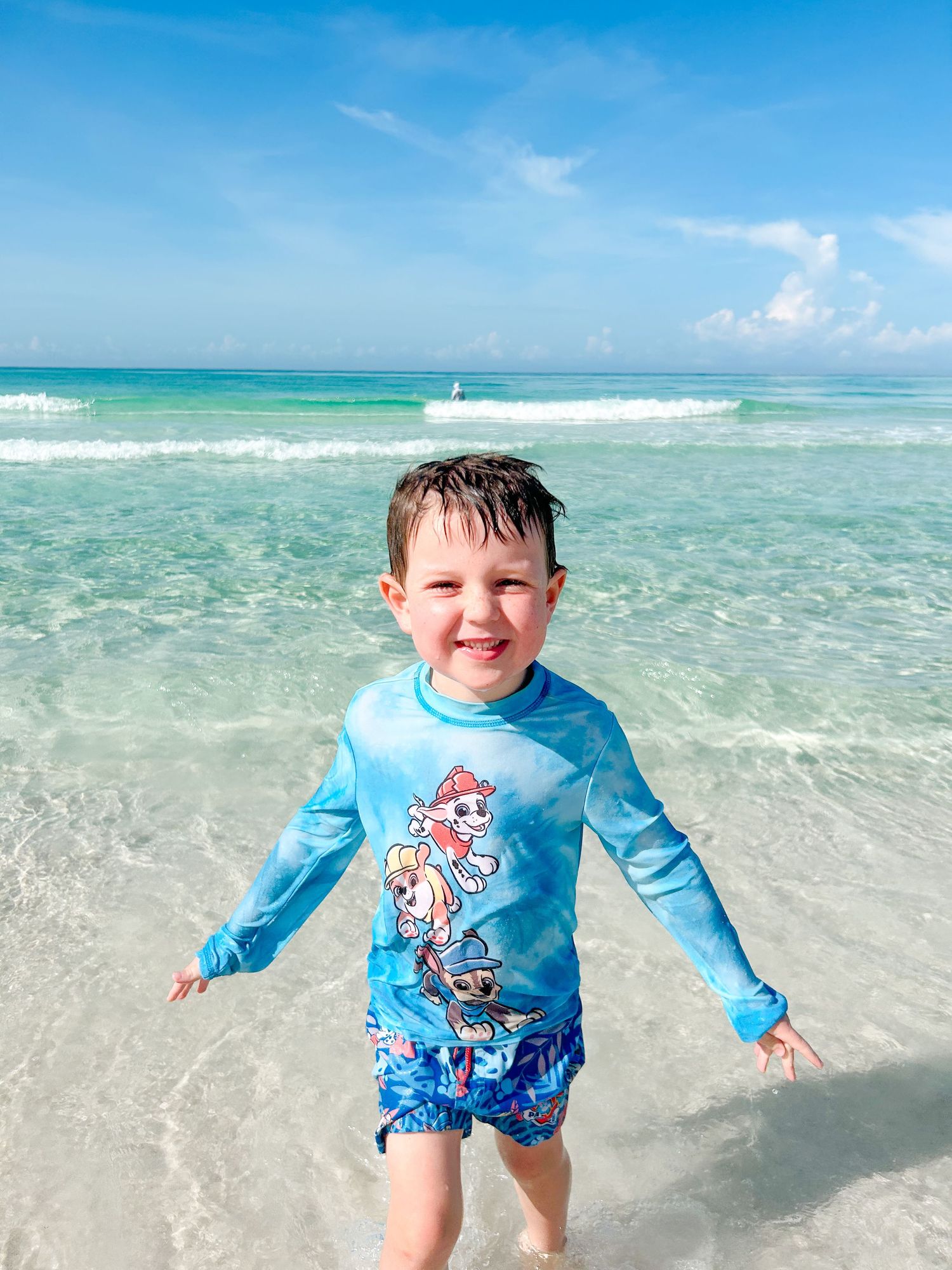 A child in a blue rash guard and swim shorts plays joyfully in the crystal clear turquoise waters of a tropical beach.