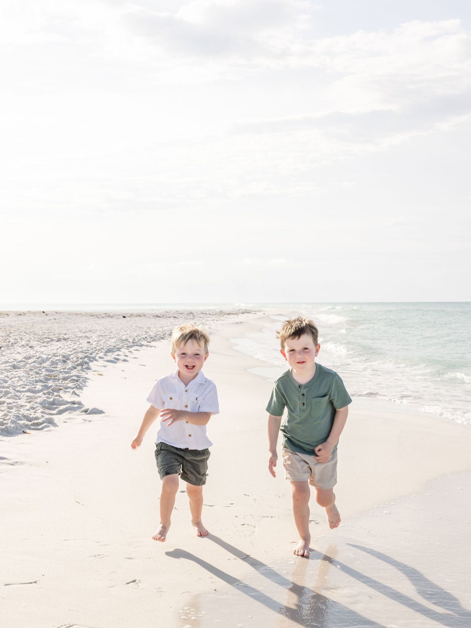 Two children run joyfully along a sunny beach shoreline with ocean waves and sandy coast in the background.